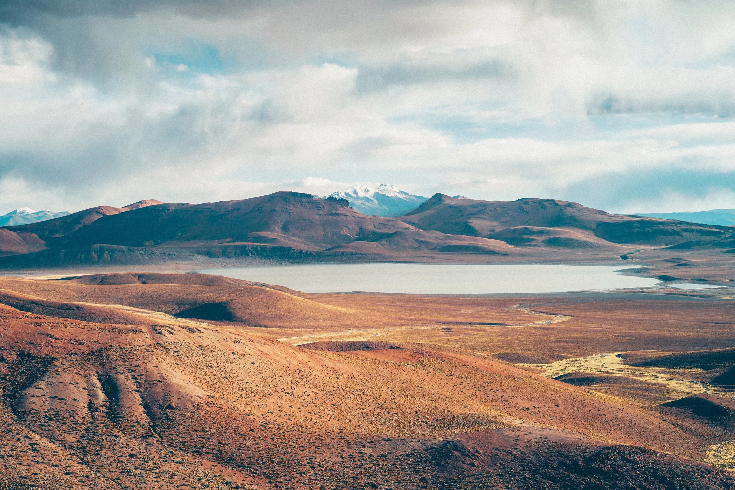 Scenic landscape of rolling hills, a large body of water, and distant snow-capped mountains under a partly cloudy sky.