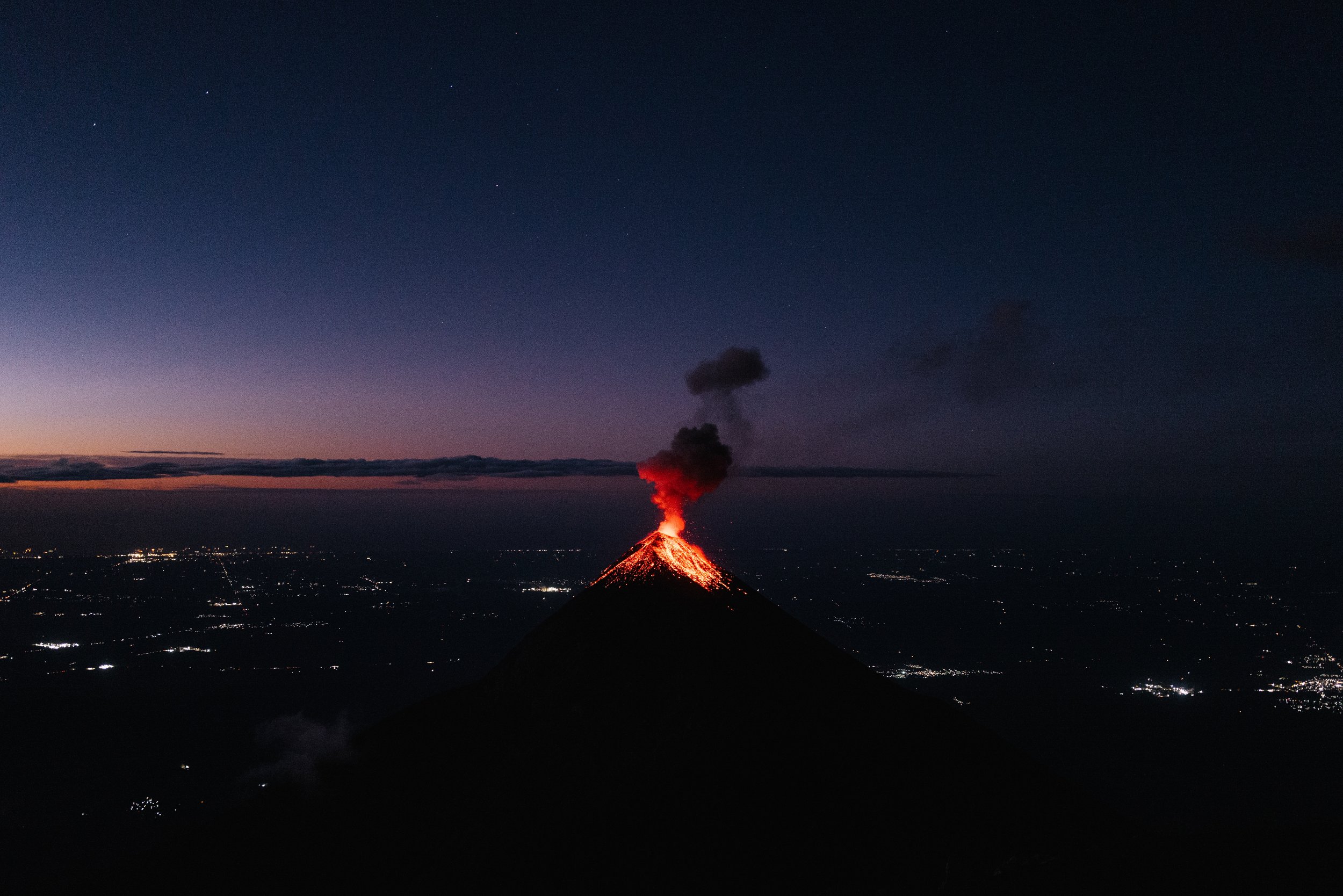 A volcano erupting at night with red lava and black smoke, surrounded by a dark landscape and a starry sky.