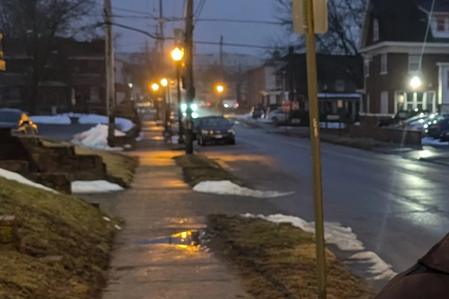 A Columbus Ohio residential street in the evening with wet pavement, patches of snow, streetlights, parked cars, and houses in the background.