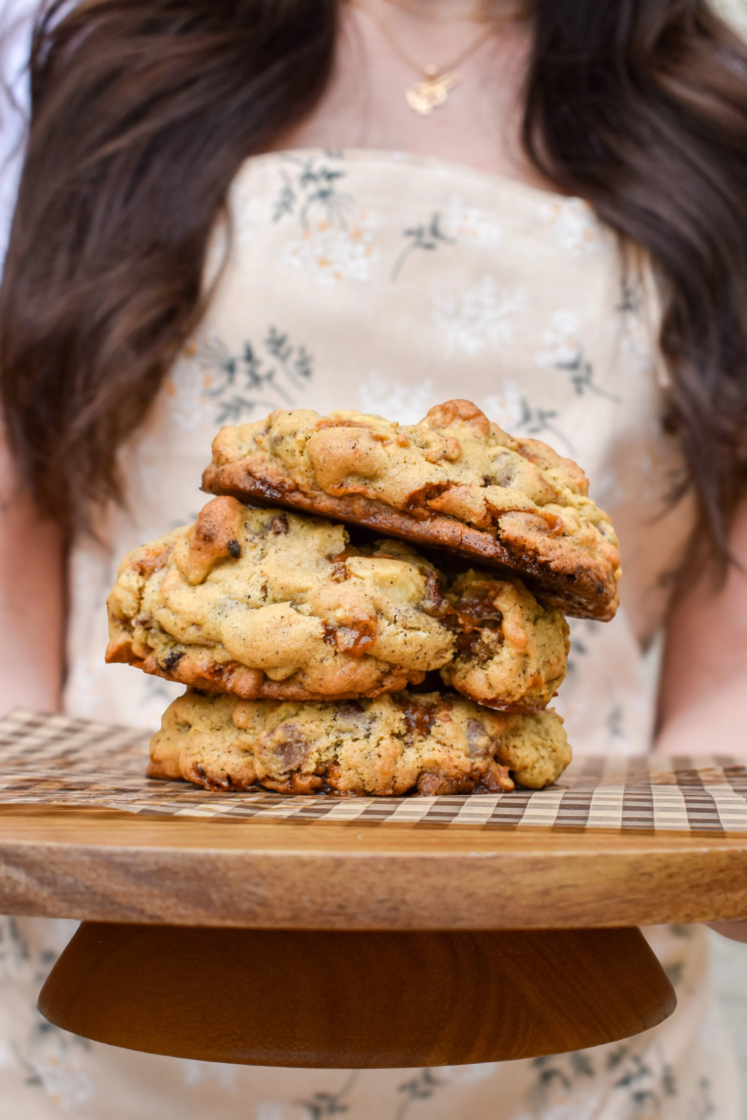 A person holding a wooden tray with three chocolate chip cookies stacked on top of each other, with part of their face and upper body visible in the background.