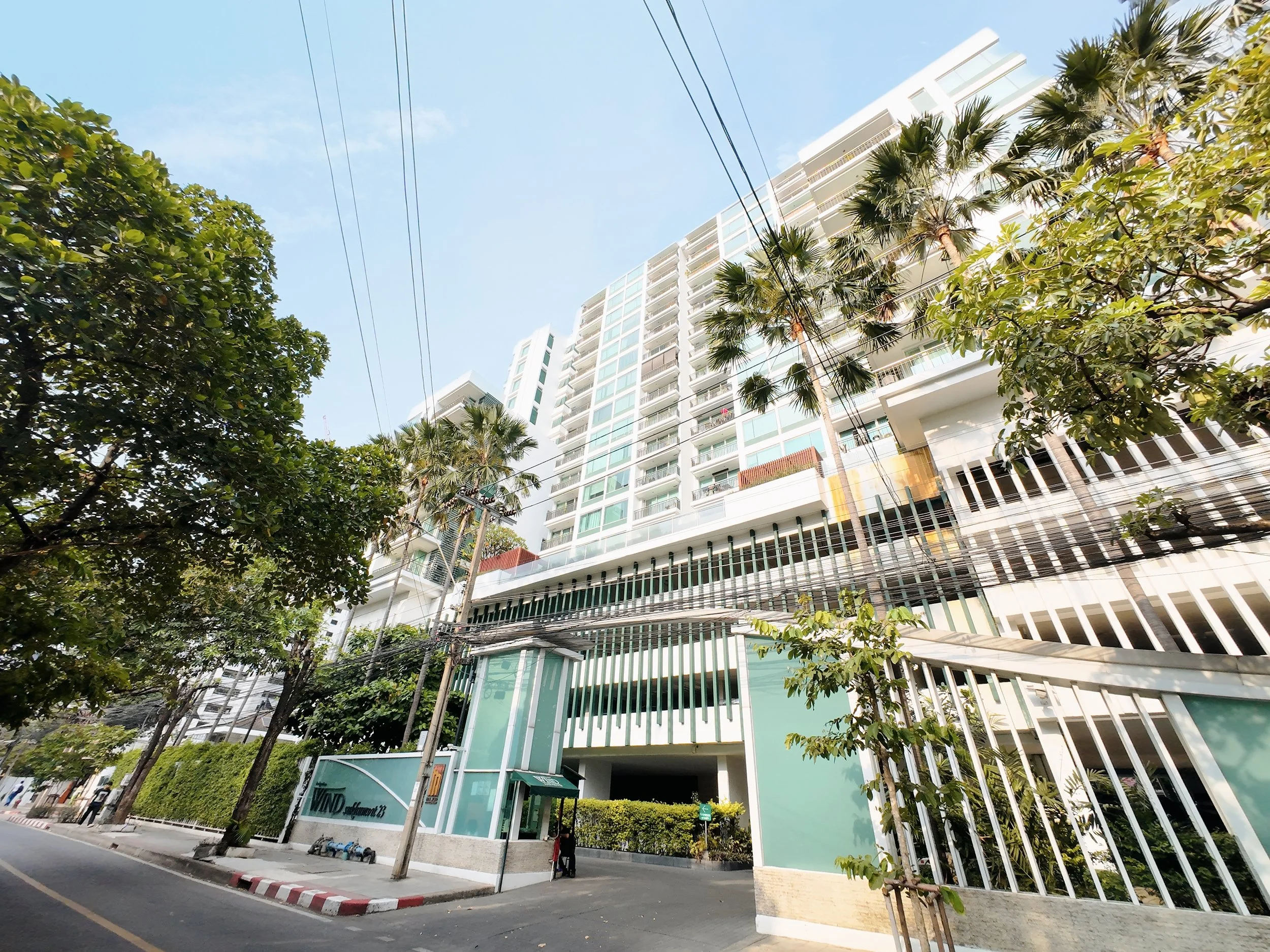 A modern high-rise building with balconies, surrounded by tall palm trees and green foliage, under a clear blue sky.