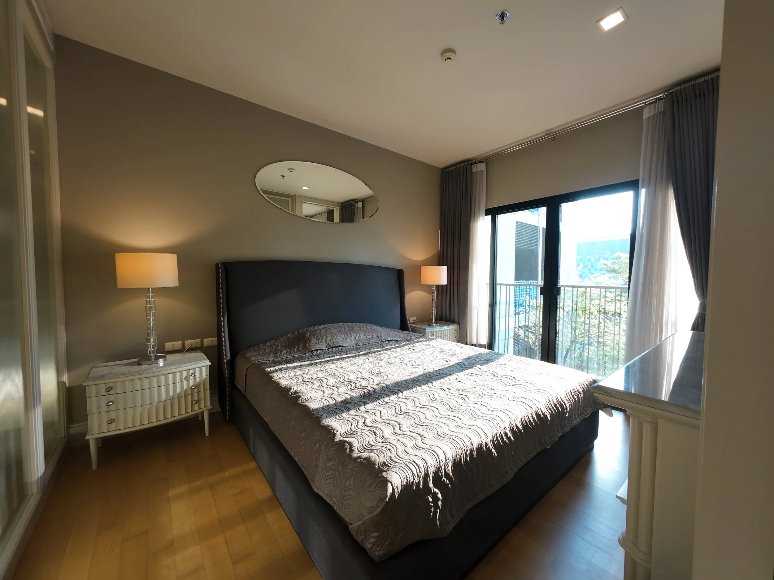 Hotel bedroom with a large bed, nightstands with lamps, a sliding glass door with curtains leading to a balcony, and a mirror on the wall.