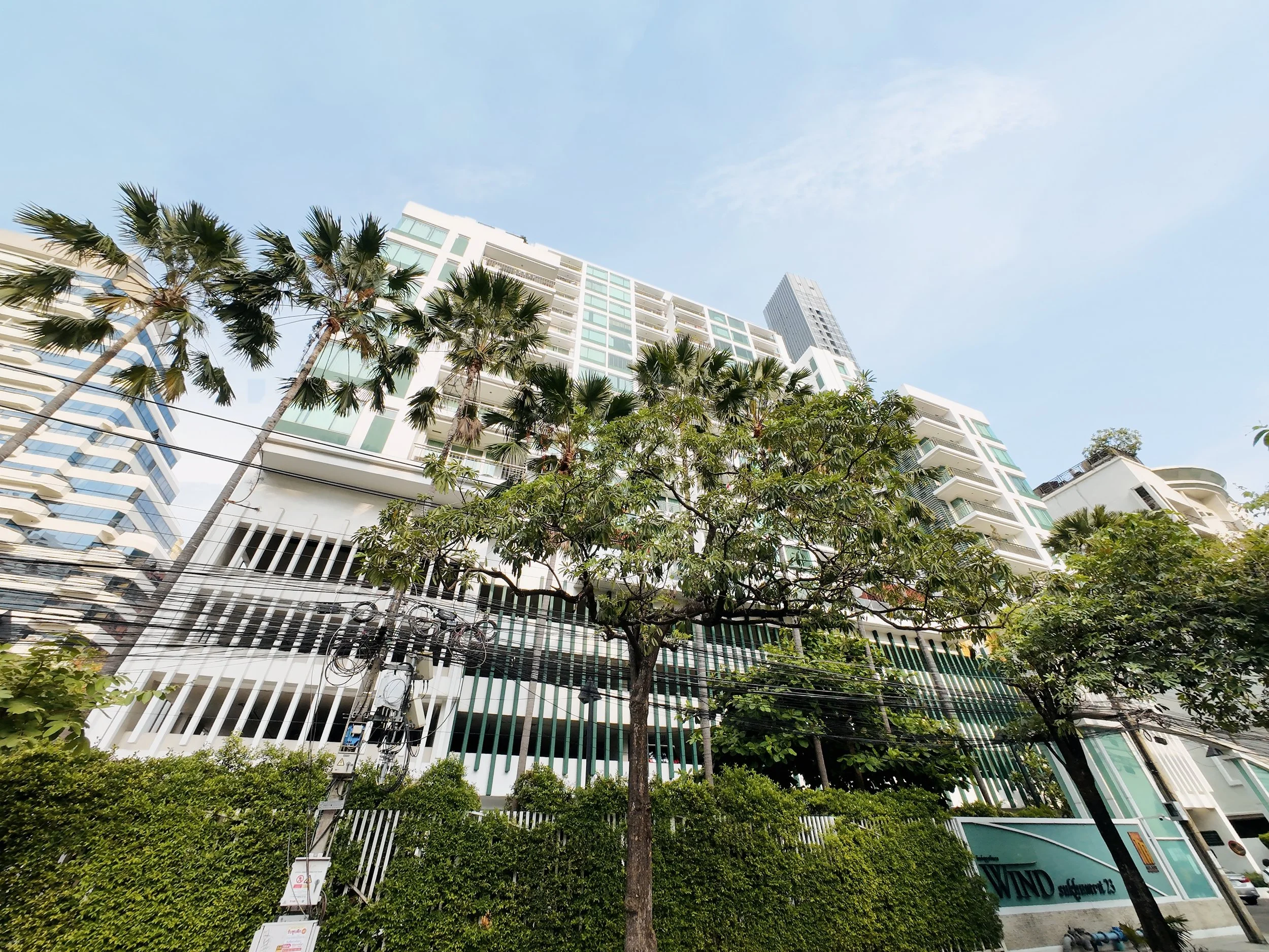 Tall modern buildings with glass windows and balconies, surrounded by lush green trees, under a clear blue sky.