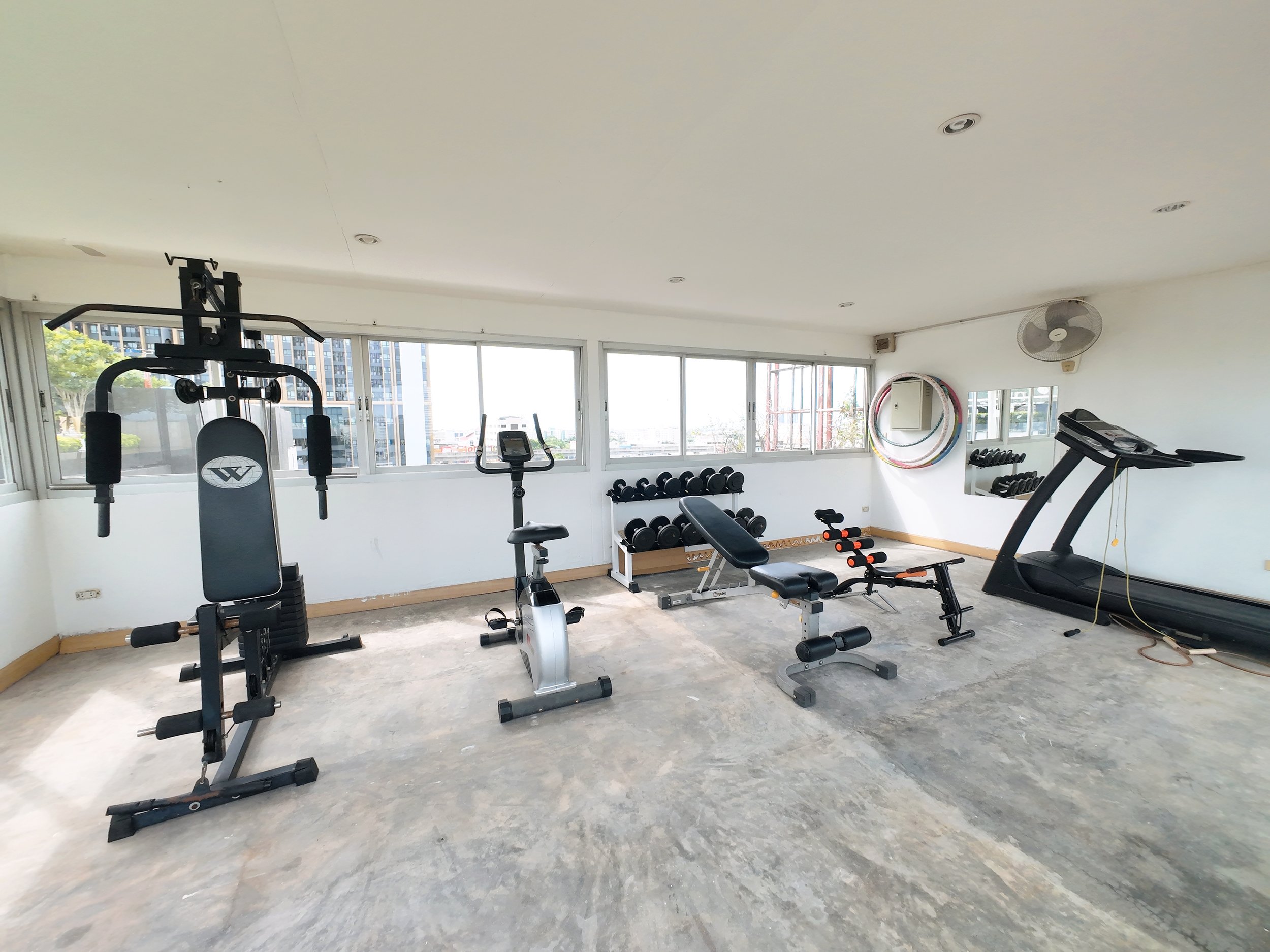 Empty gym room with various exercise equipment including a weight machine, stationary bike, incline bench, small dumbbells, and a treadmill. Large windows overlook a cityscape.