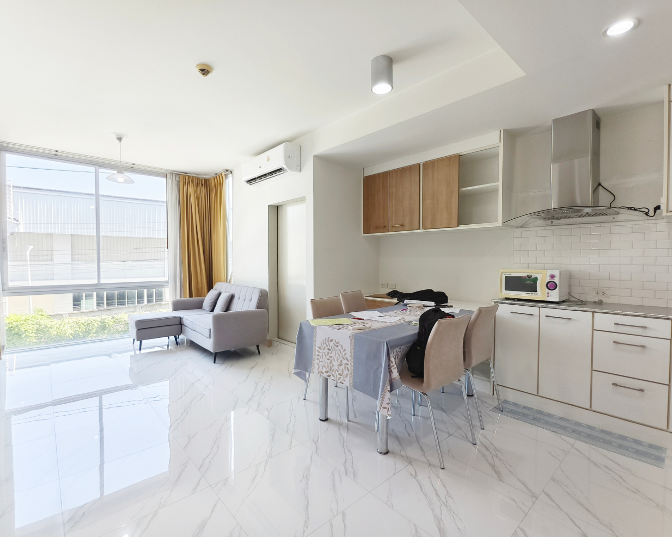 Modern living room with large window, white marble floor, gray sofa, dining table with beige chairs, kitchen area with white cabinets and microwave, yellow curtains, and ceiling lights.