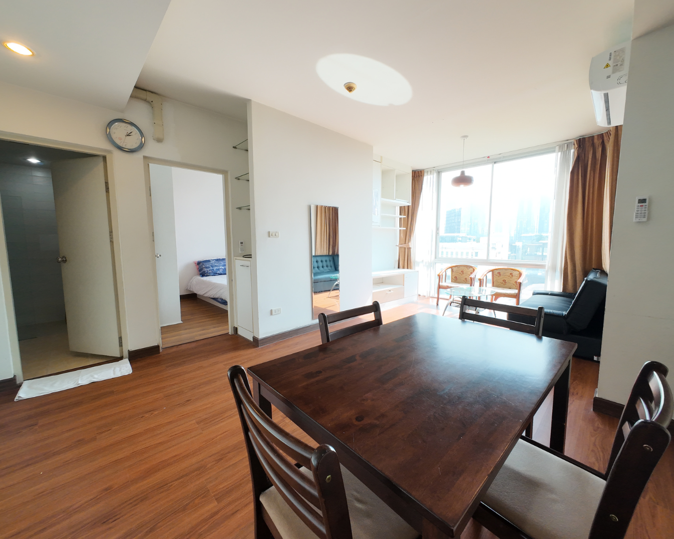 Interior of an apartment with a wooden dining table, chairs, a living area with large windows, curtains, and a view of city buildings outside.