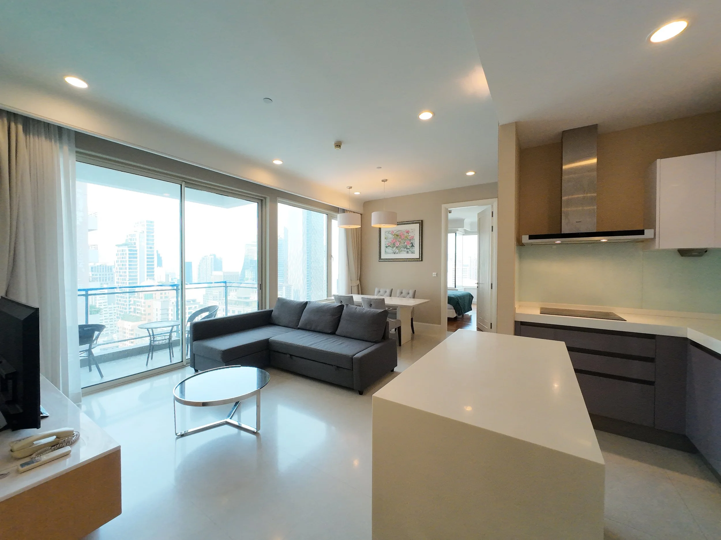 Living room with a gray sofa, glass coffee table, large windows with city view, white ceiling with recessed lighting, and a kitchen area with white and black cabinets.