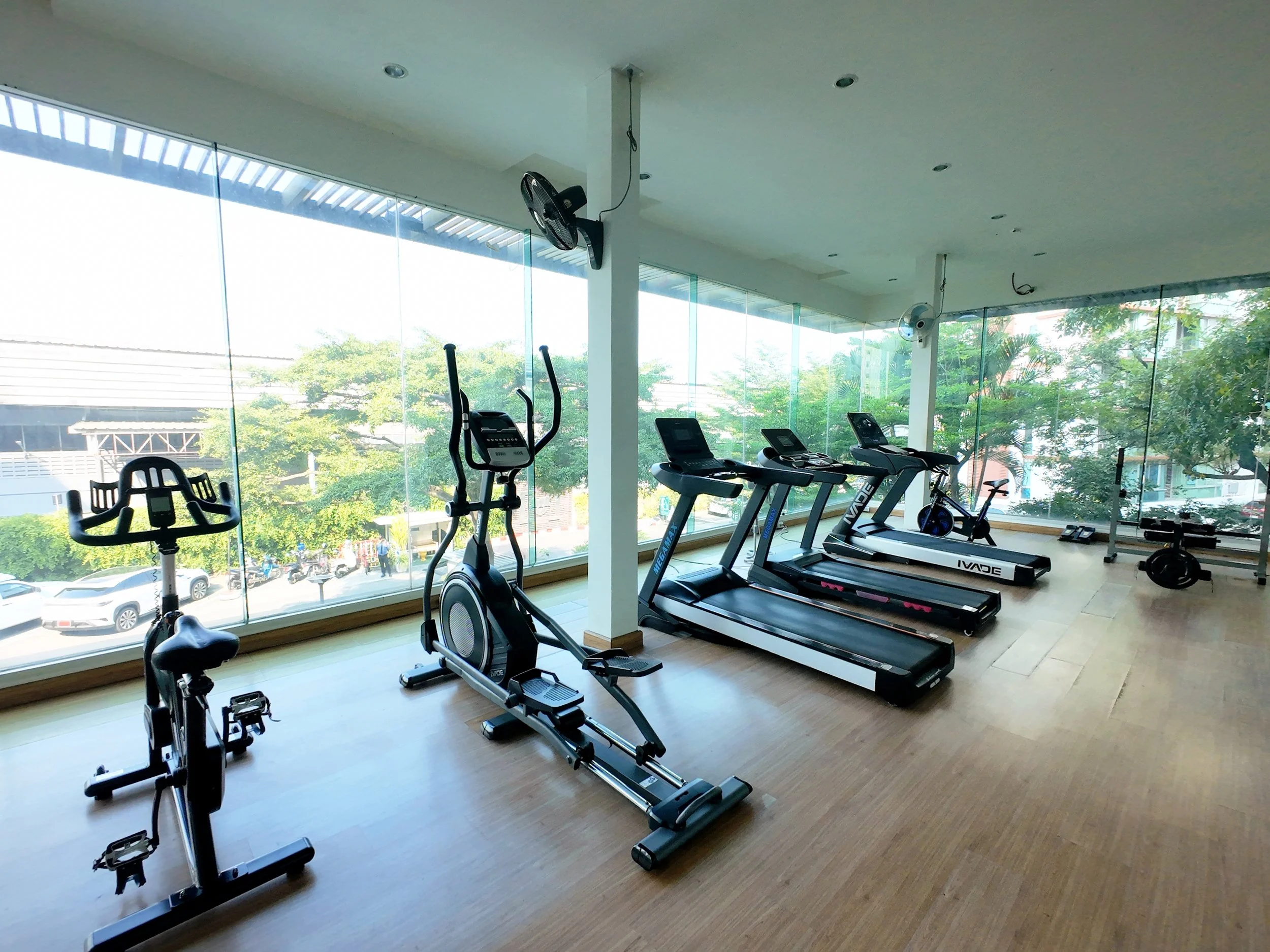Gym with large windows, several exercise machines including treadmills, stationary bike, and elliptical equipment, with trees visible outside.