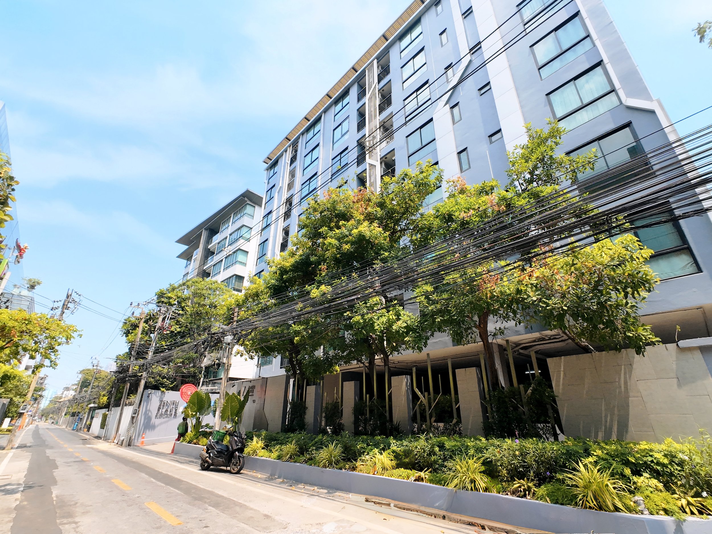 Street view of a modern high-rise building with trees, power lines, and a scooter parked on the side of the road.