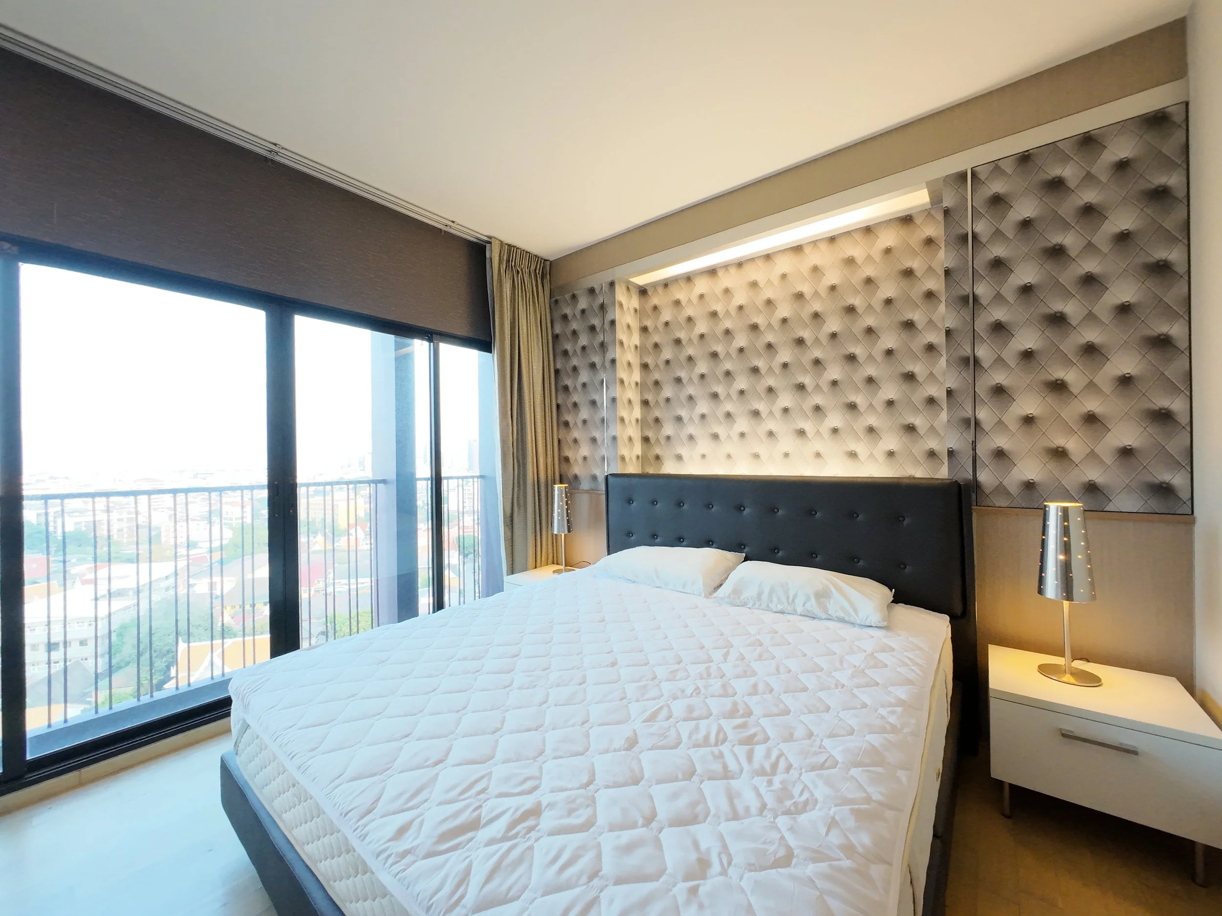Modern bedroom with large window, sliding glass door with city view, bed with black headboard, bedside lamps, and textured wall panels.