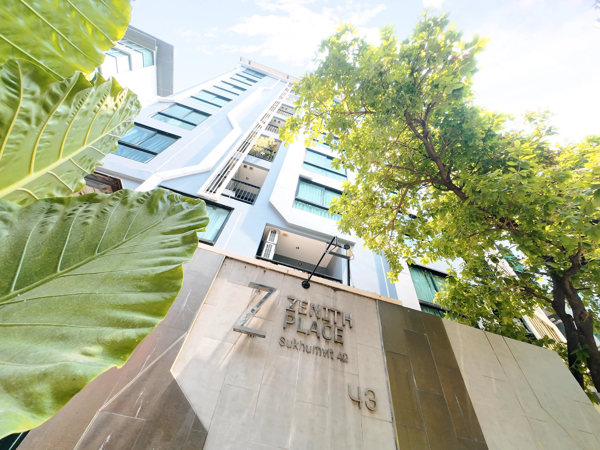 View of a modern white building with large windows, surrounded by green trees and large jungle leaves. The photo is taken from ground level looking up towards the sky. There is a sign at the bottom with the text 'EQUIPMENT PLACE Sukhumvit 42' visible.