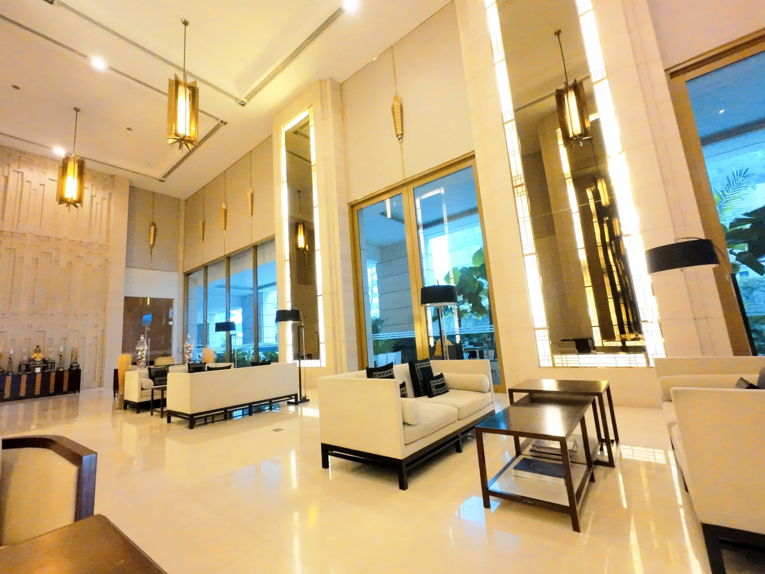 The image shows a spacious modern lobby area with high ceilings, large windows, and contemporary furniture, including white sofas, black chairs, and wooden tables. Elegant hanging pendant lights with a gold finish illuminate the space, and decorative plants are visible outside through the windows.