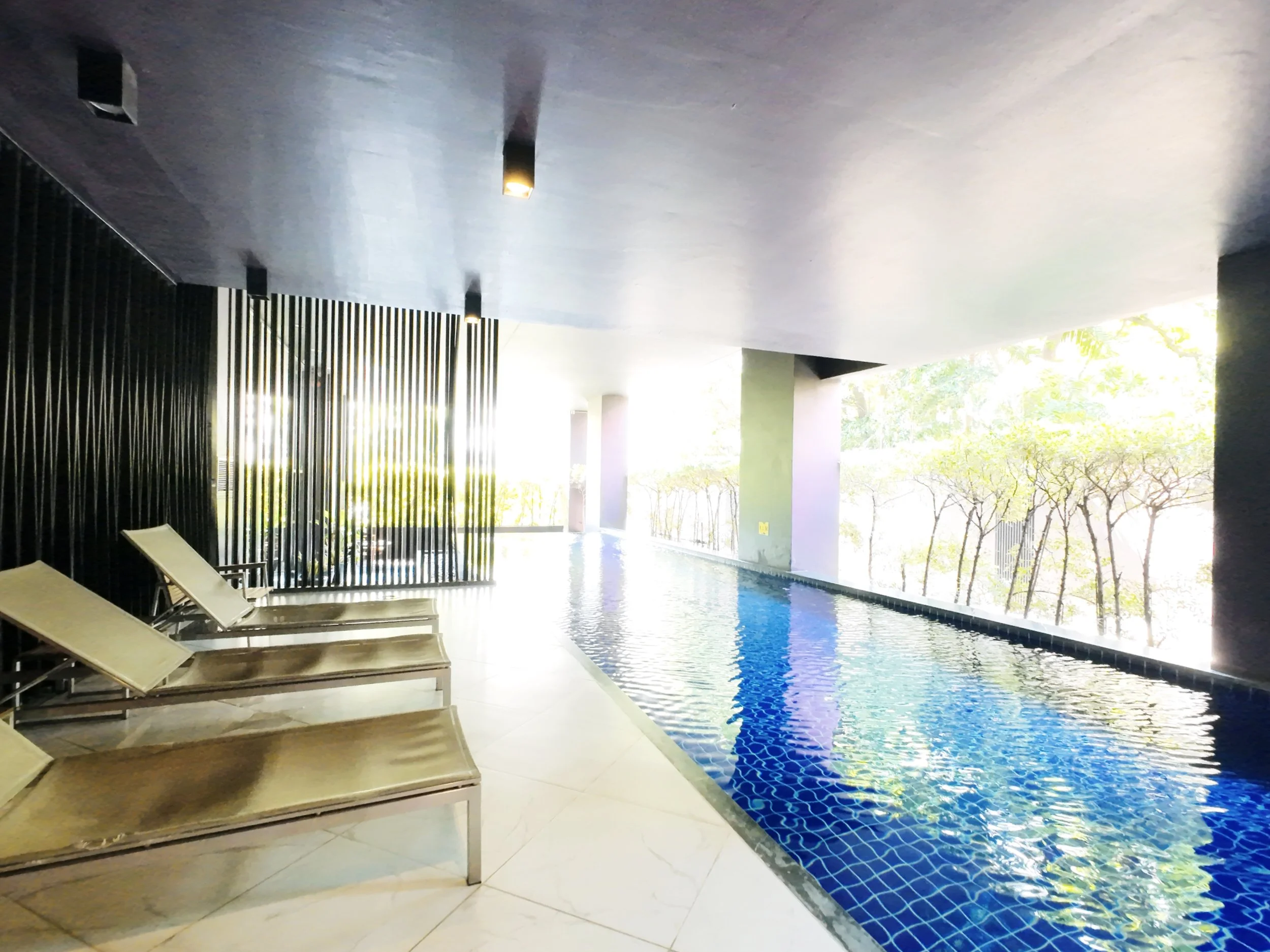 Indoor swimming pool area with four beige lounge chairs, black ceiling, and large windows with trees outside.