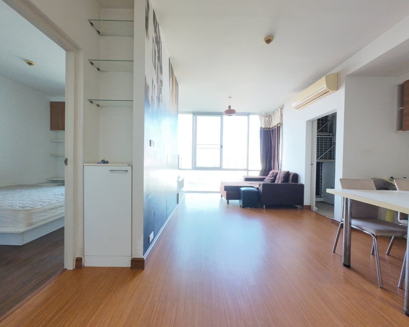 Interior of an apartment with wooden flooring, a living room with a sofa, large windows with curtains, and a dining area with a table and chairs.