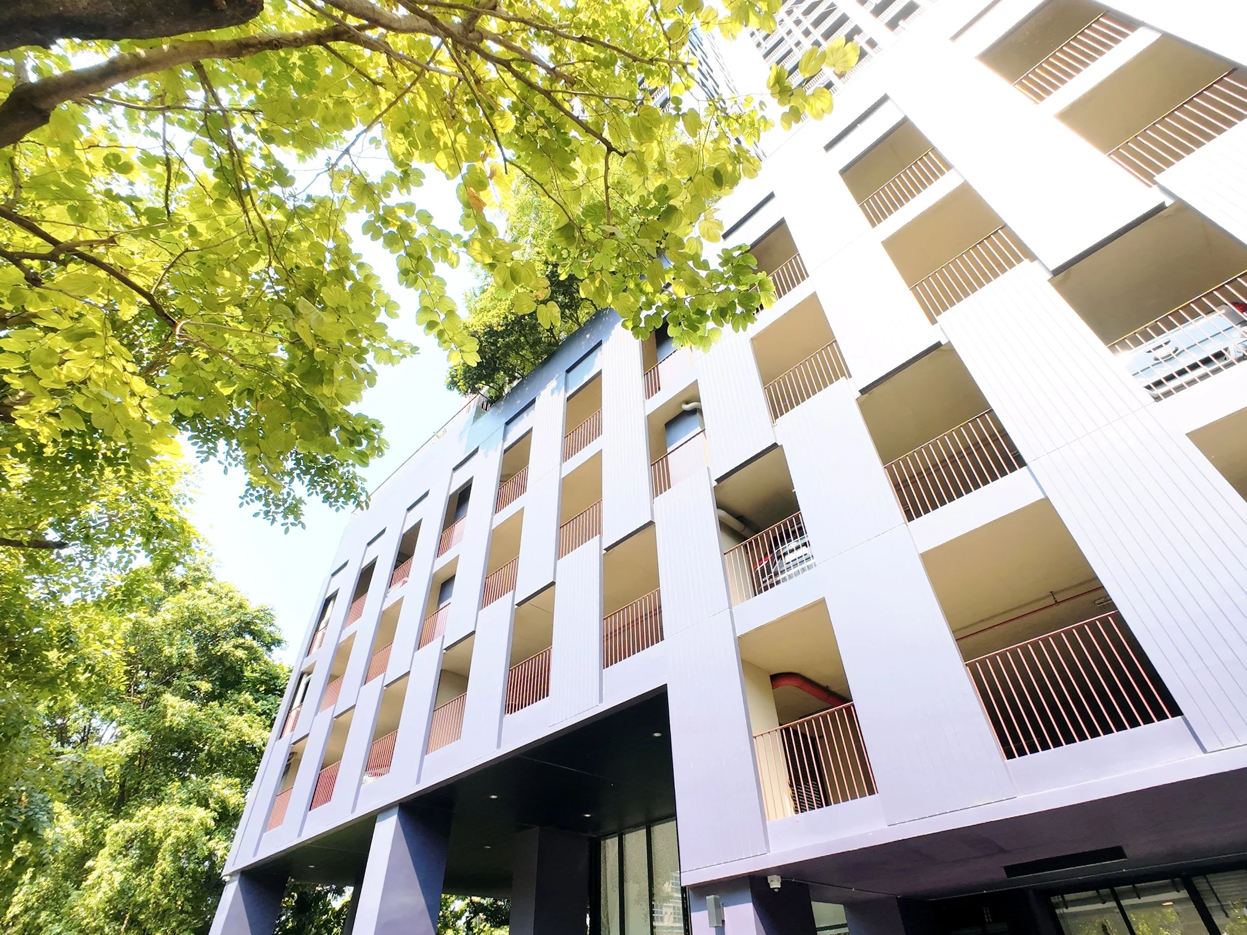Low-angle view of a modern multi-story apartment building with white vertical panels and red balcony railings, surrounded by green trees against a bright sky.