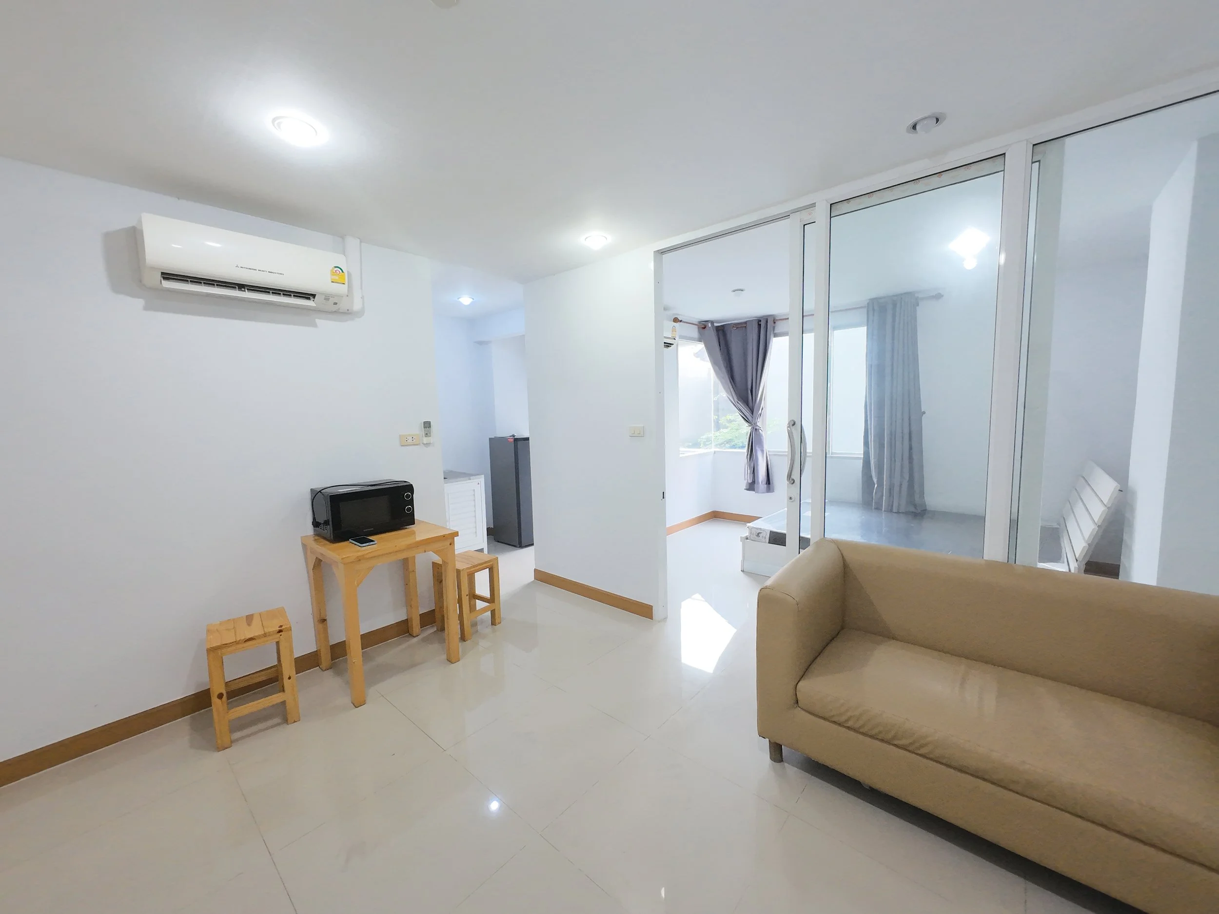 Living room with beige sofa, glass sliding doors leading to a bedroom with a bed and curtains, a small wooden table with a microwave, two small wooden stools, white walls, tiled floor, air conditioner, and some ceiling lights.