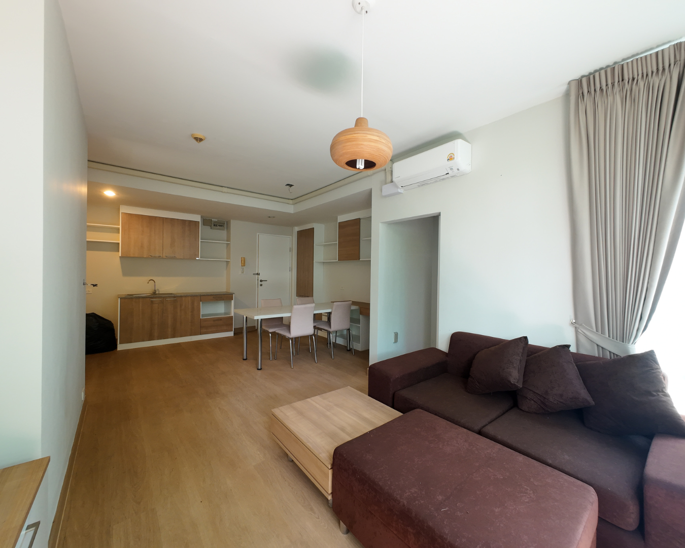 Living room and kitchenette with wooden furniture, a dining table with four chairs, brown sofa with cushions, large window with curtains, air conditioning unit on the wall, wooden ceiling light fixture.