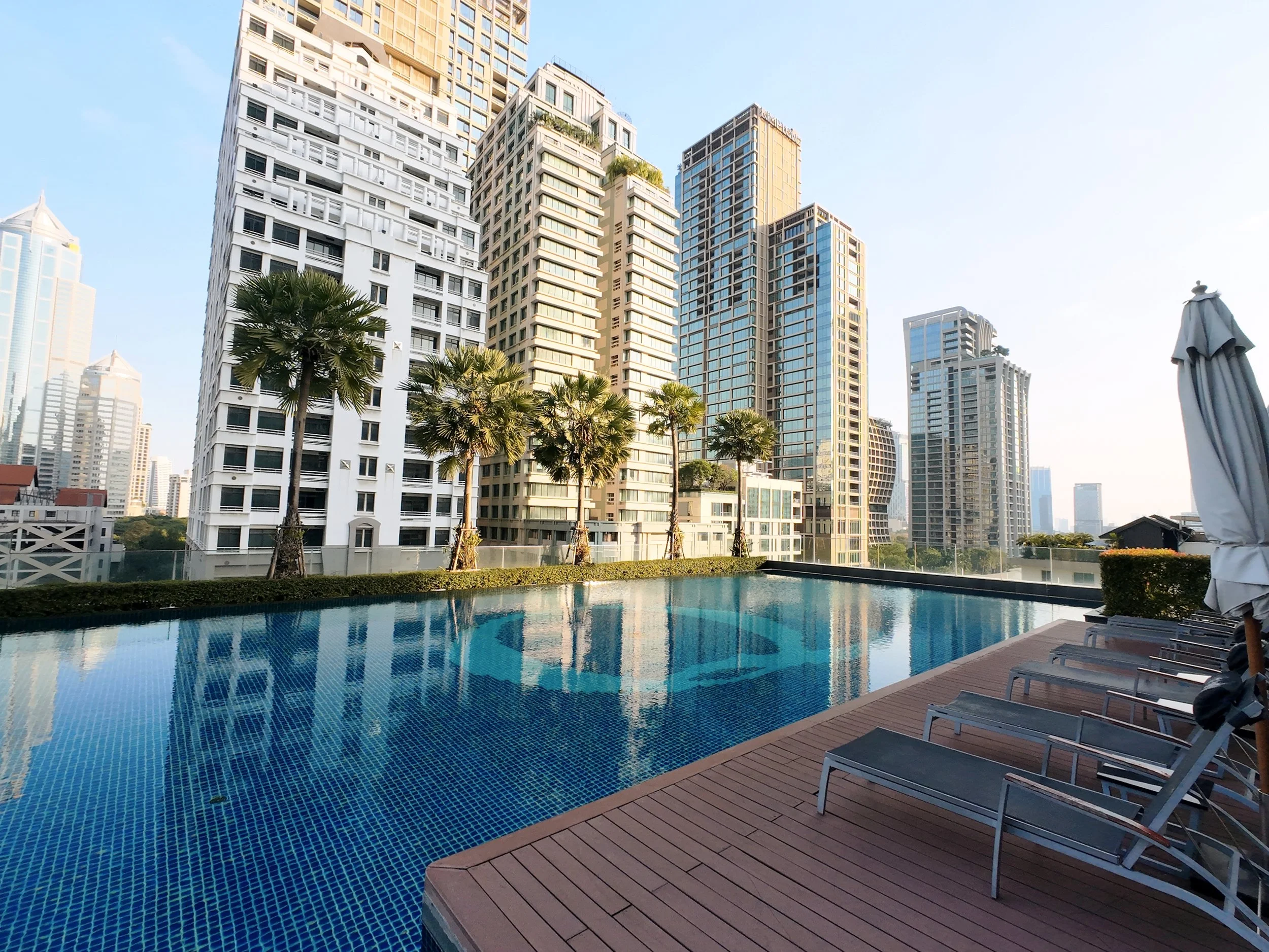A rooftop pool area with a blue tiled swimming pool, poolside lounge chairs, and a city skyline with tall buildings in the background.