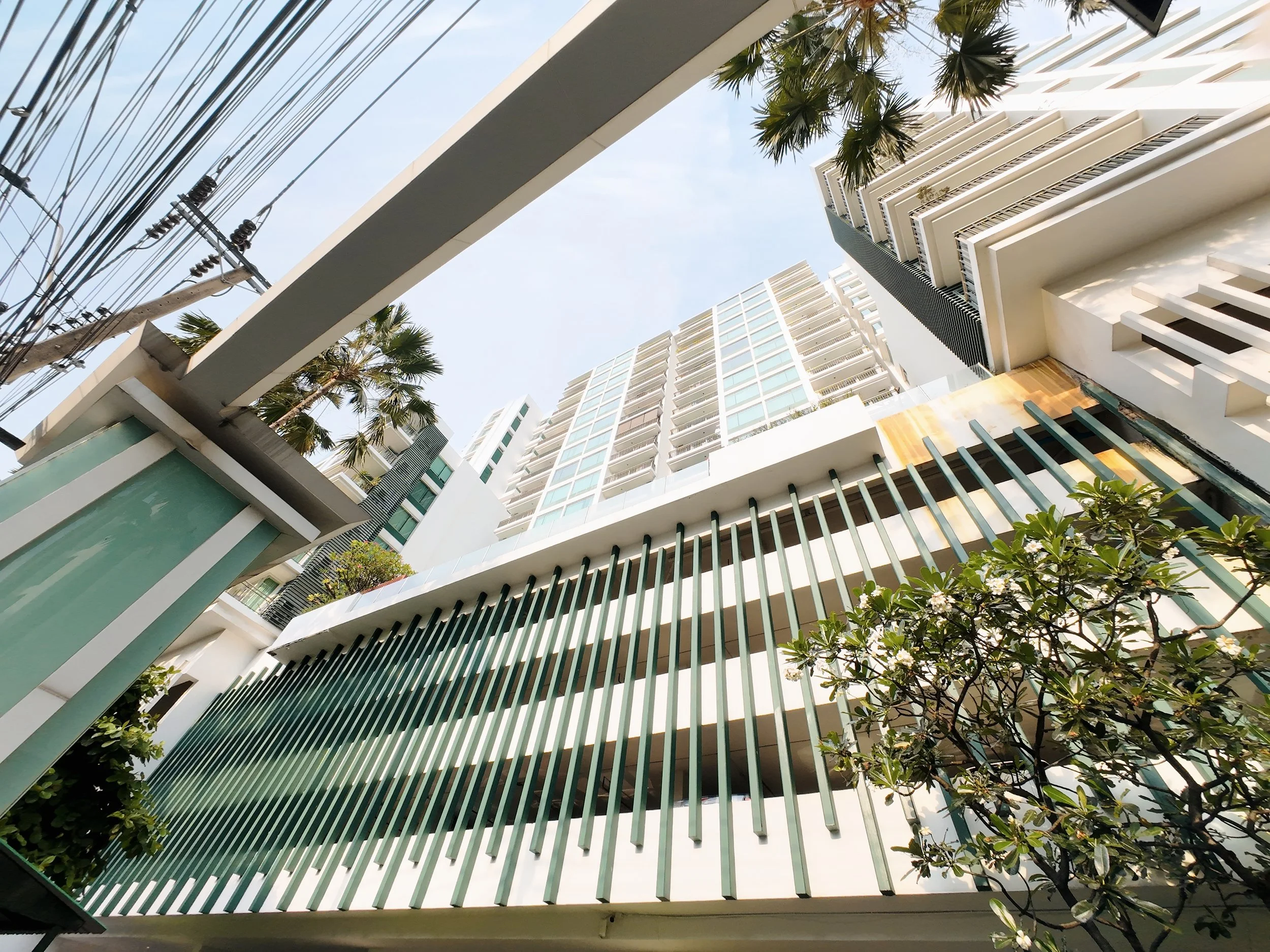 Looking up at modern white high-rise buildings with glass windows, palm trees, and a blue sky with some clouds.