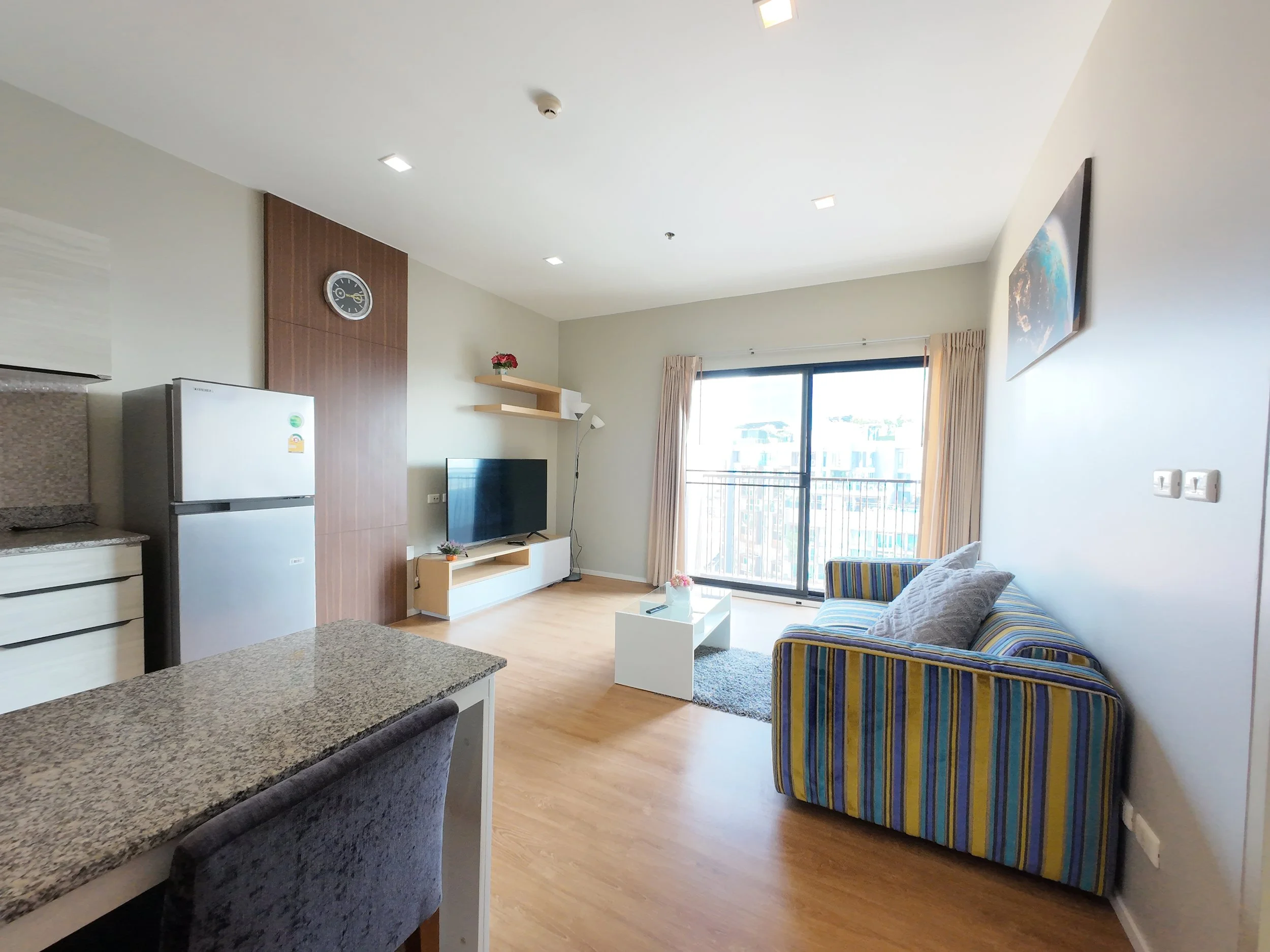 Living room with a striped sofa, coffee table, television, sliding glass door with curtains, refrigerator, kitchen counter, and a wall clock.