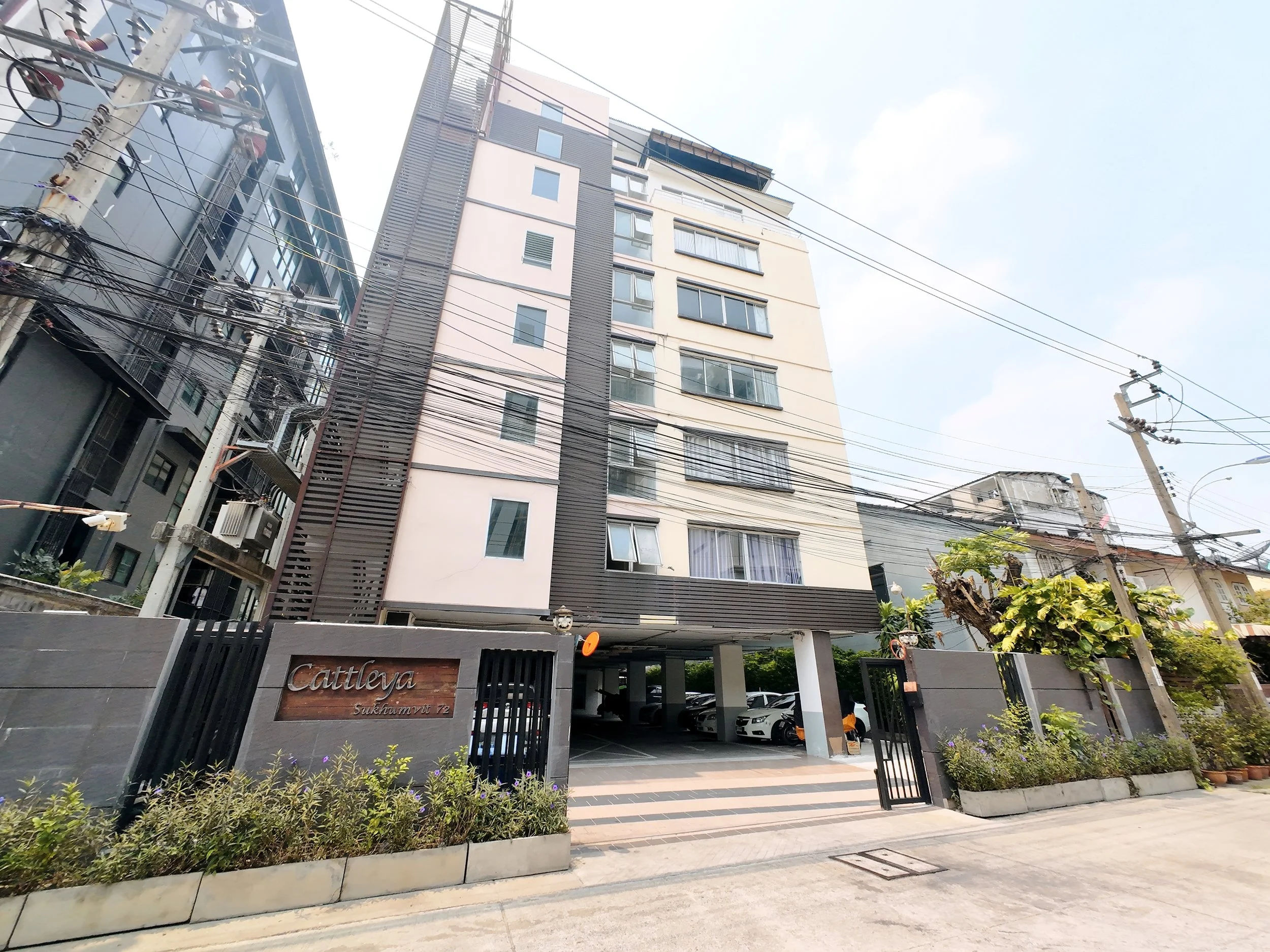 Multistory residential building with parking area under the building, the building is labeled 'Cattleya Sukhumvit 72' at the entrance. There are electric wires across the street and some potted plants with flowers outside.