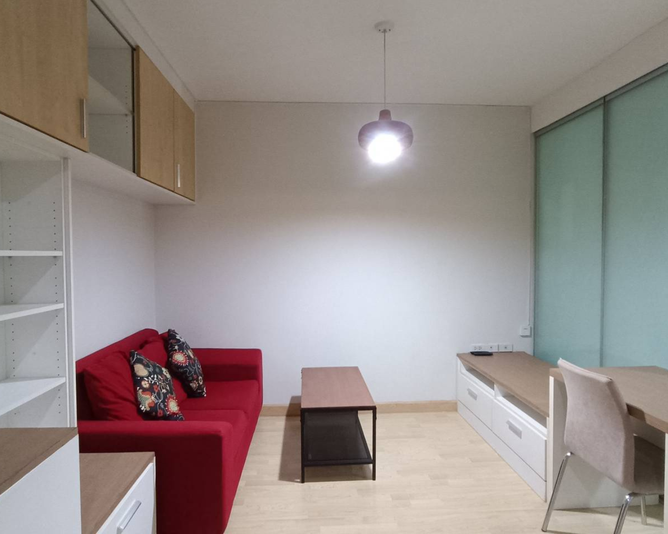 Living room with red sofa, wooden coffee table, white shelving, built-in storage, table, and chair, with a hanging ceiling light, light wood flooring, and a frosted glass sliding door.