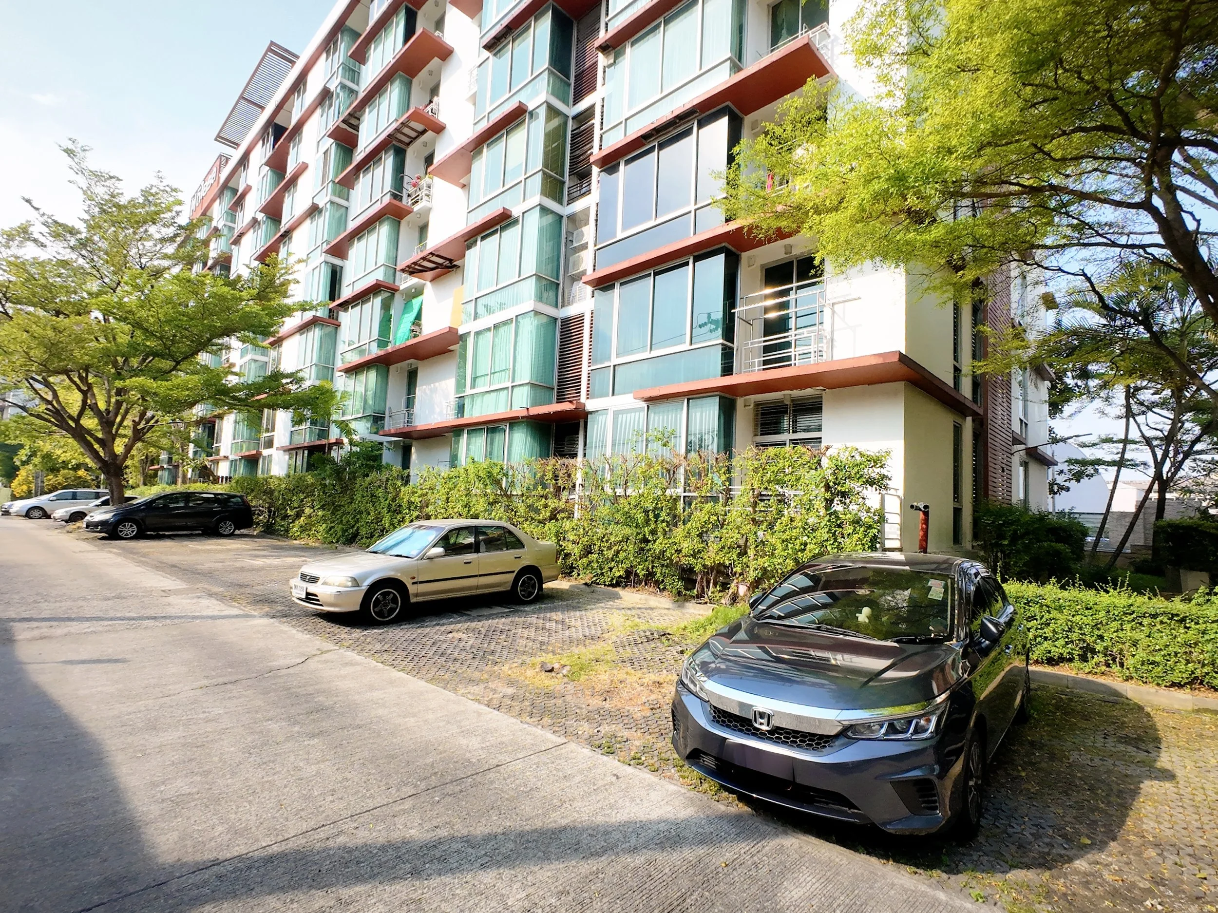 Apartment building with glass balconies, parked cars including a Honda, trees, and shrubbery in a sunny urban setting.