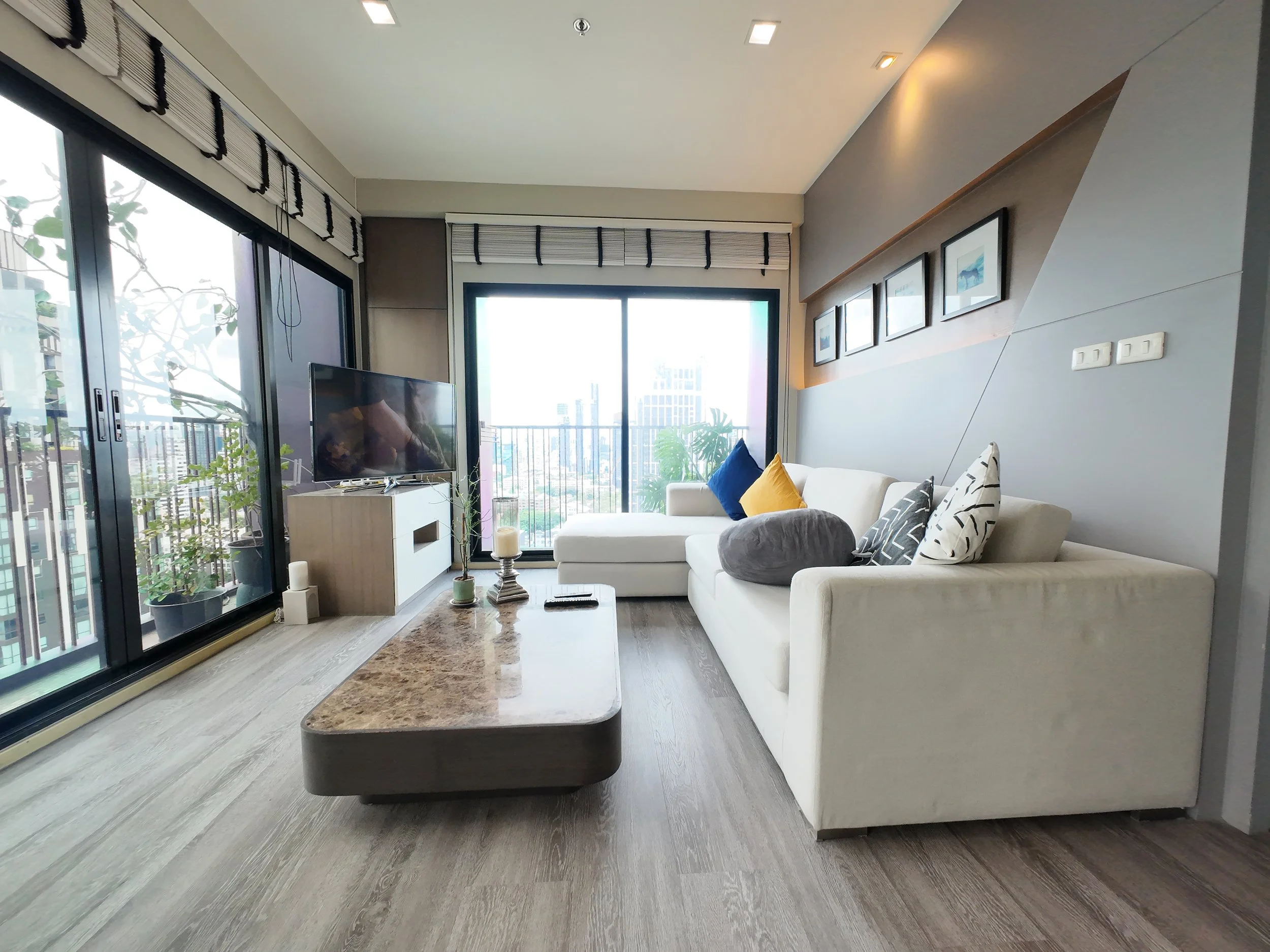 Modern living room with sliding glass doors opening to a balcony with city views. The room features a white sofa with various pillows, a marble coffee table, a flat-screen TV on a white console, and framed artwork on the wall. There are indoor plants near the balcony.