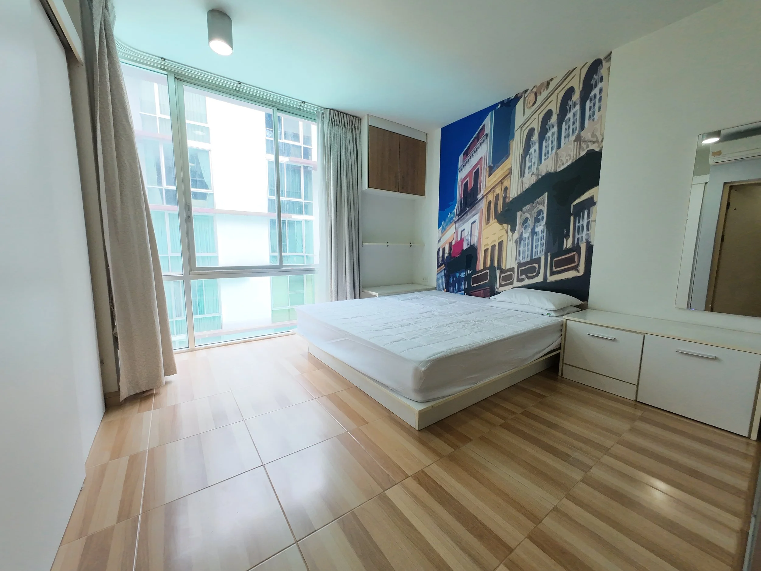 A bedroom with a large window, beige curtains, a bed with white sheets, a colorful mural of a cityscape on the wall, and wooden flooring.