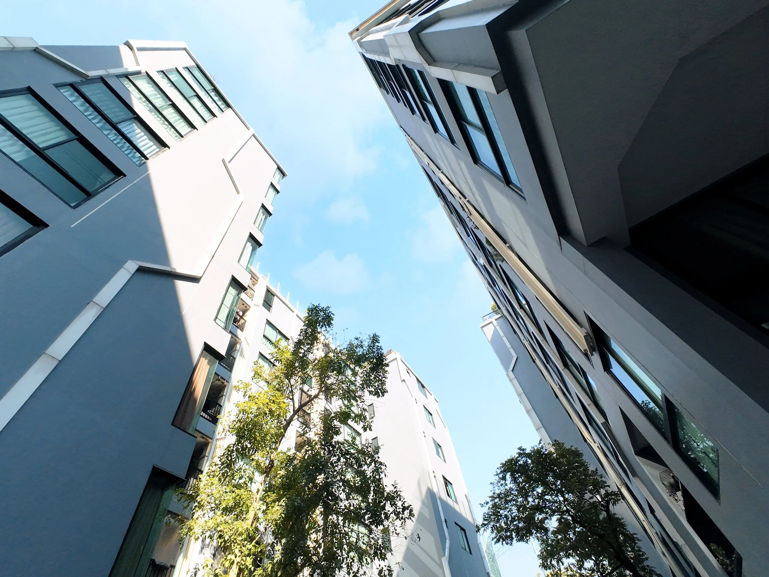 Looking up at modern white apartment buildings with glass windows, surrounded by green trees, under a blue sky with some clouds.