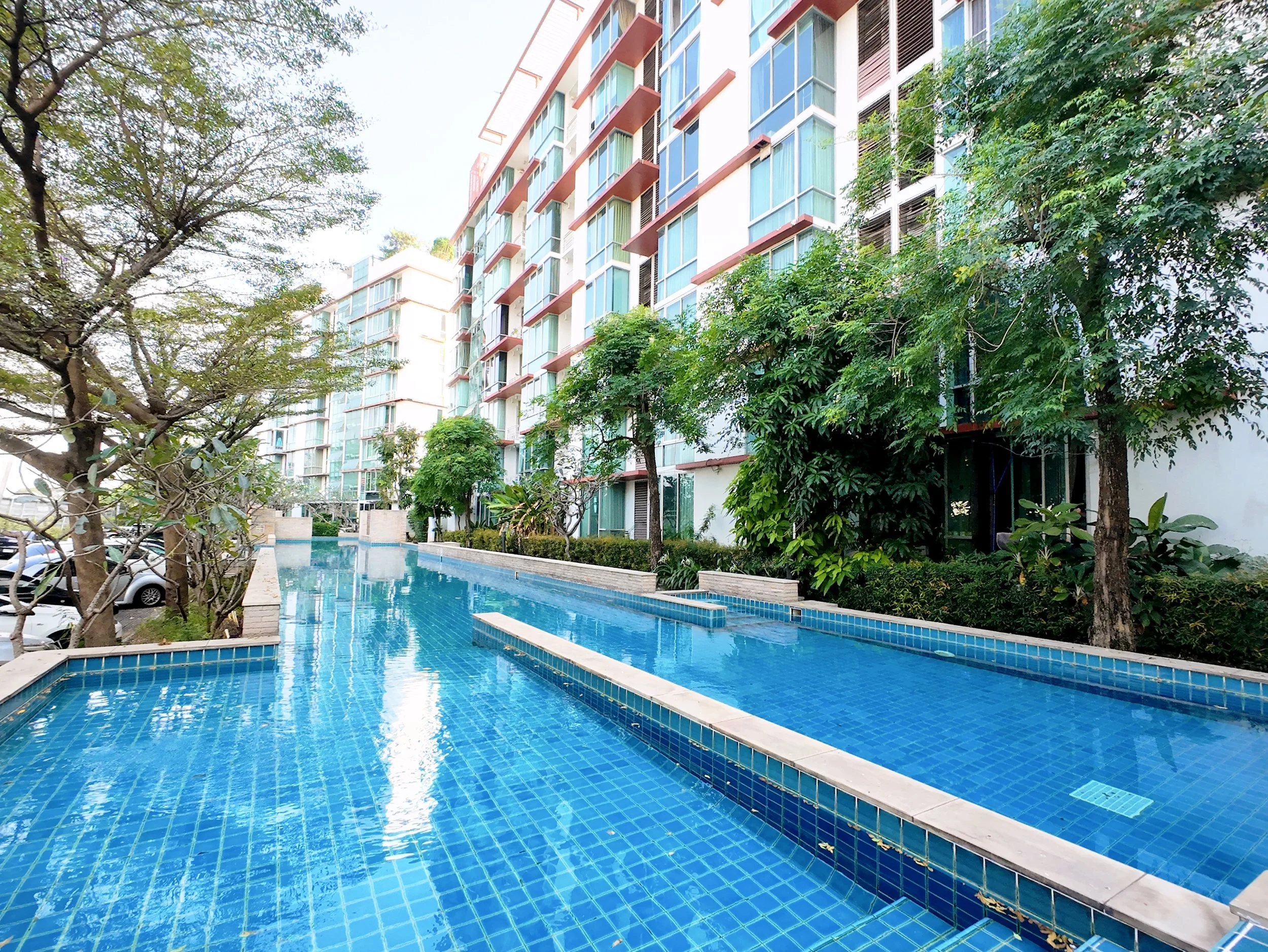 Exterior view of a modern apartment complex with a swimming pool, surrounded by trees and parking lot