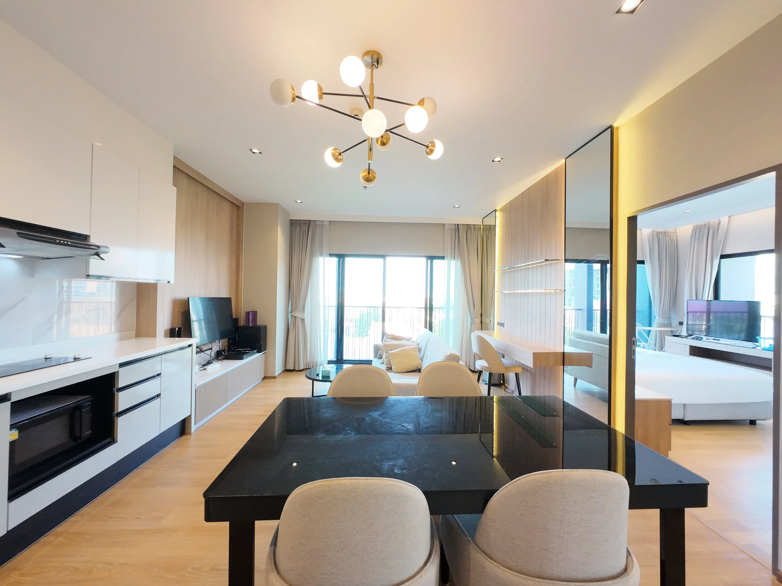 Modern living room and kitchen area with black dining table, beige chairs, white sofa, flat-screen TV, wooden flooring, and large windows with curtains.