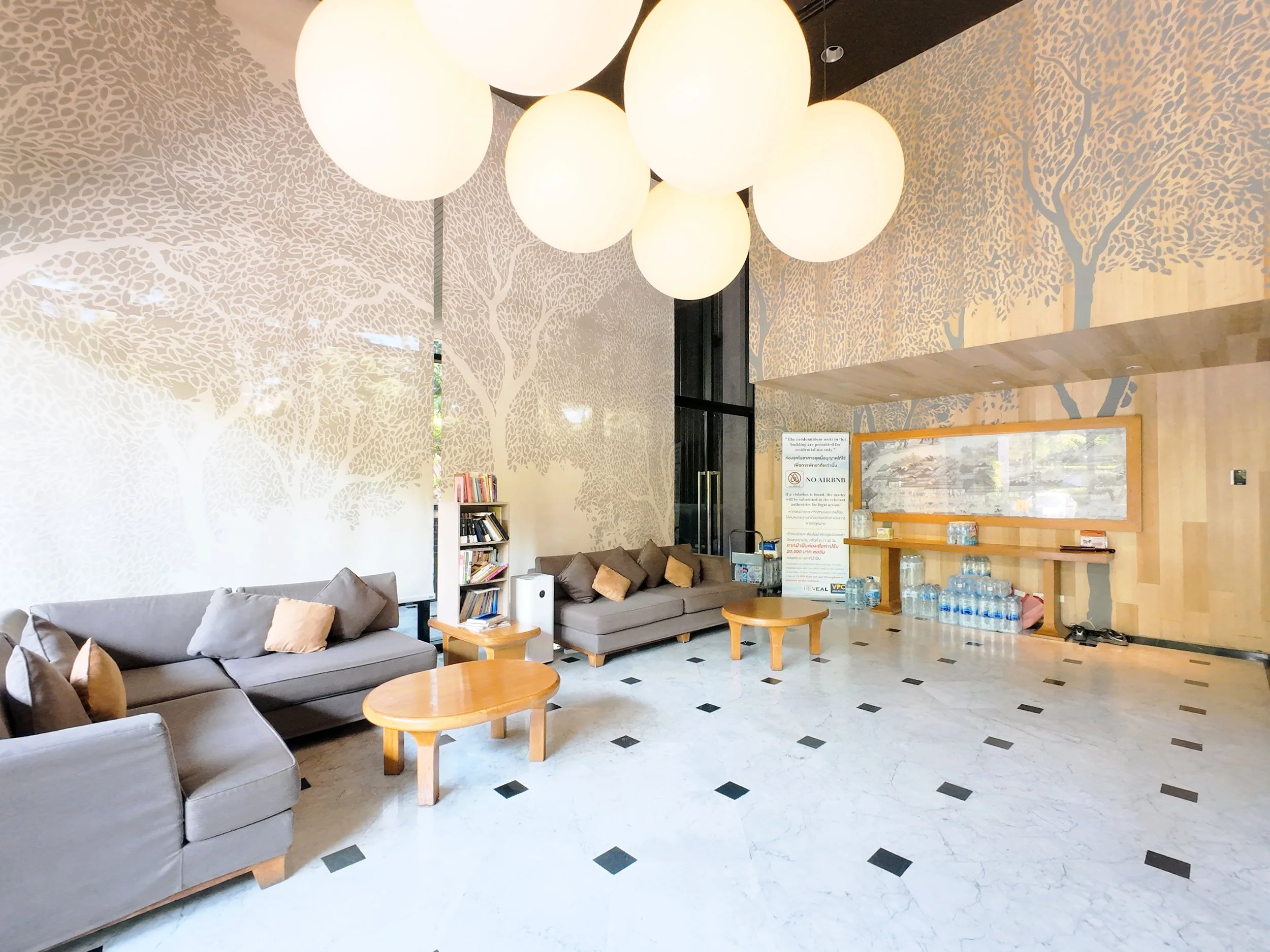 Lobby area with beige sofas, small wooden tables, a bookshelf, water bottles, and wall murals of trees, with a large hanging light fixture.
