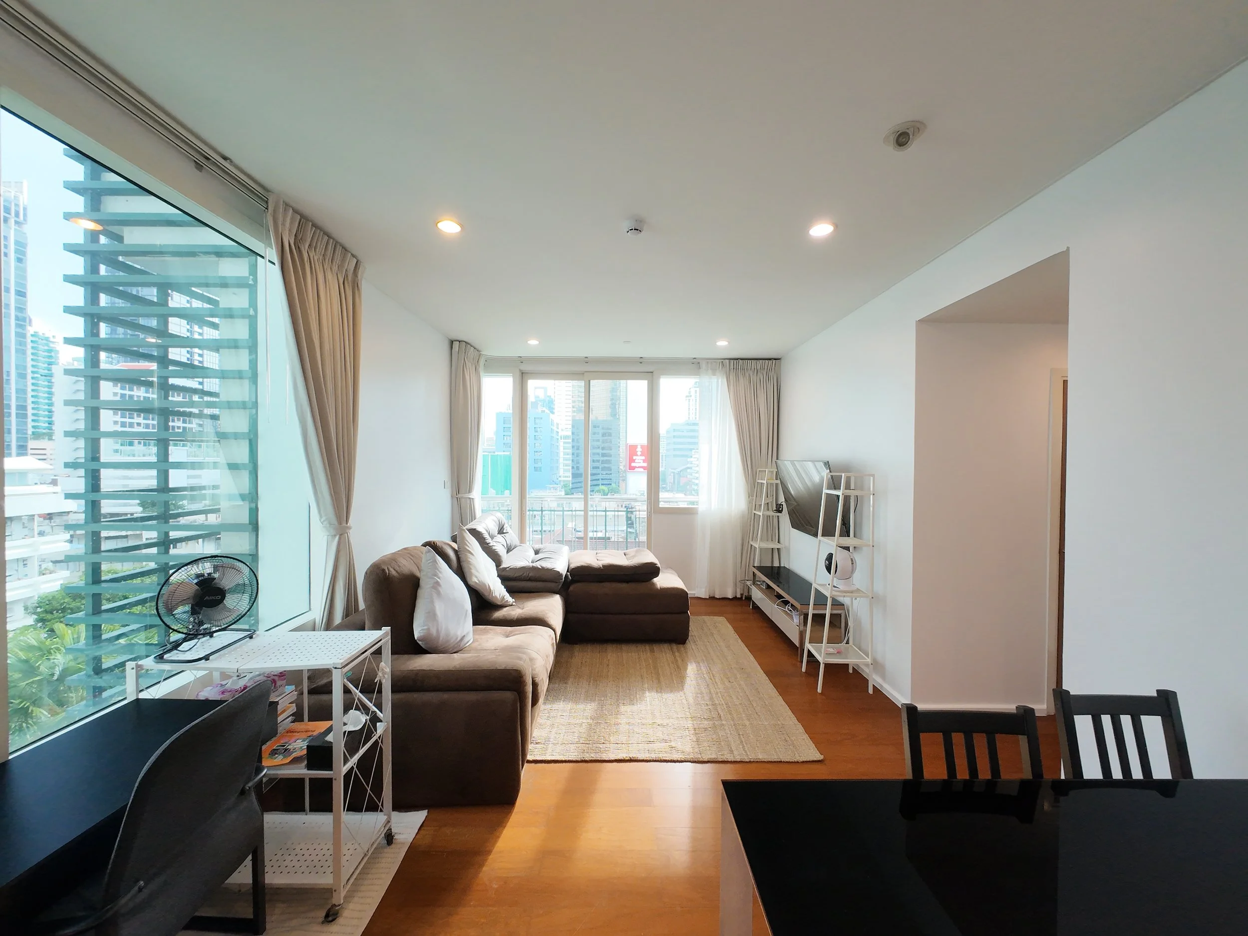 Living room with a brown sectional sofa, white side table, black dining table with chairs, and white shelving unit with a TV, in a high-rise apartment with large windows and city view.