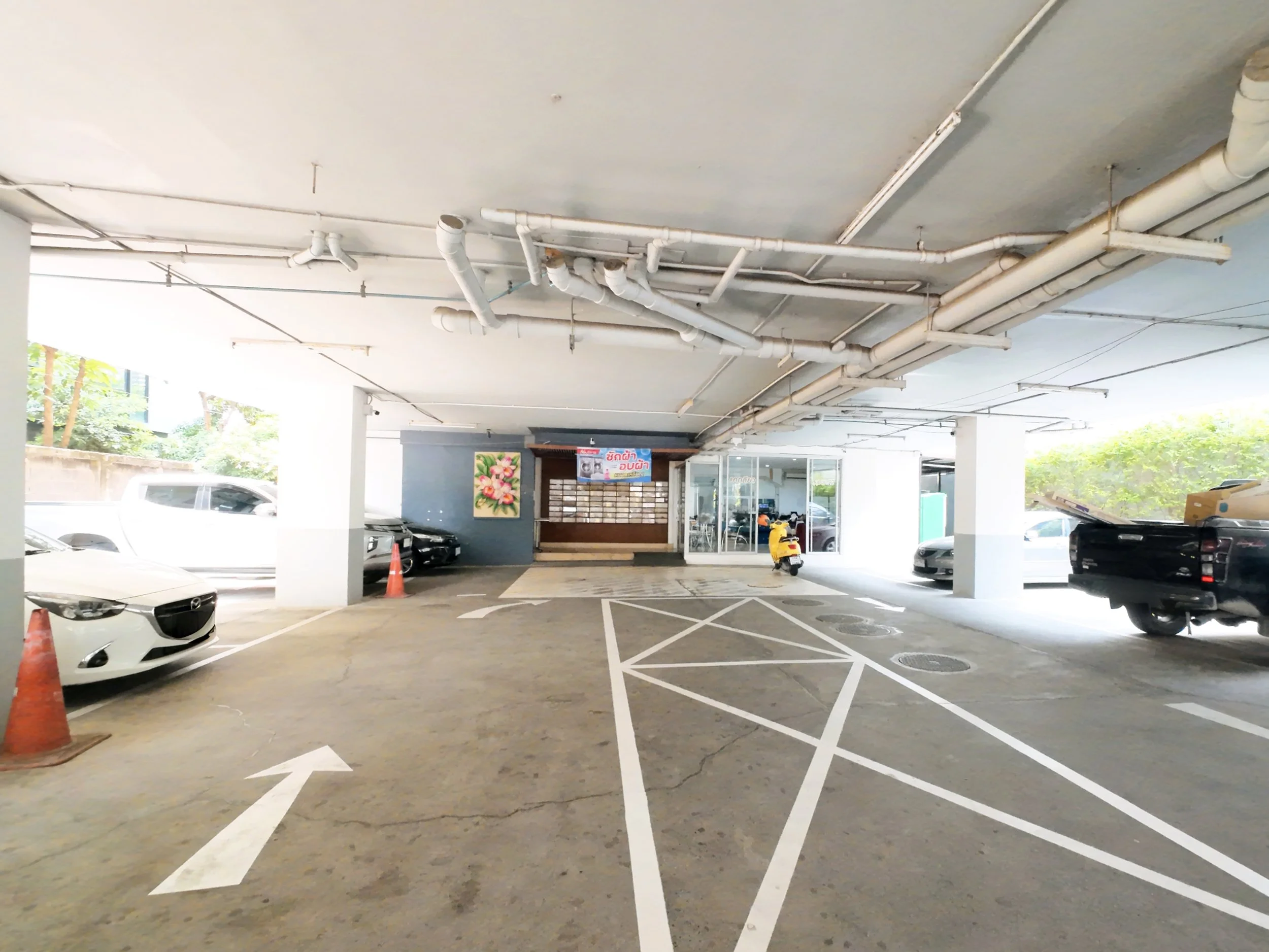 Underground parking lot with cars parked on the sides, white arrows and markings on the concrete floor, and a small yellow scooter parked near the entrance. There are pipes on the ceiling and an entrance to a building at the back.