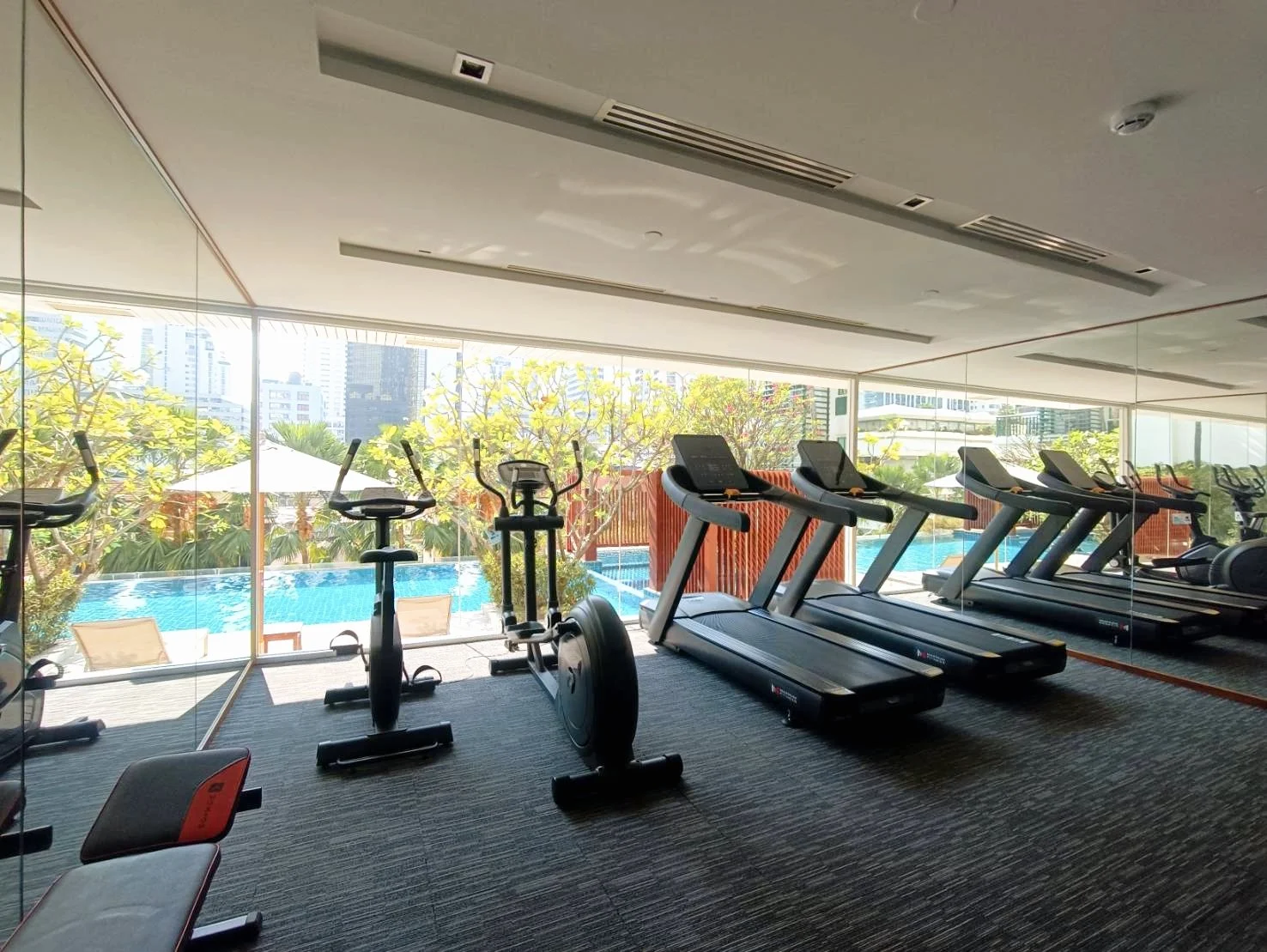Indoor gym with treadmills, exercise bikes, and weight bench, overlooking outdoor pool and city skyline through large glass windows.