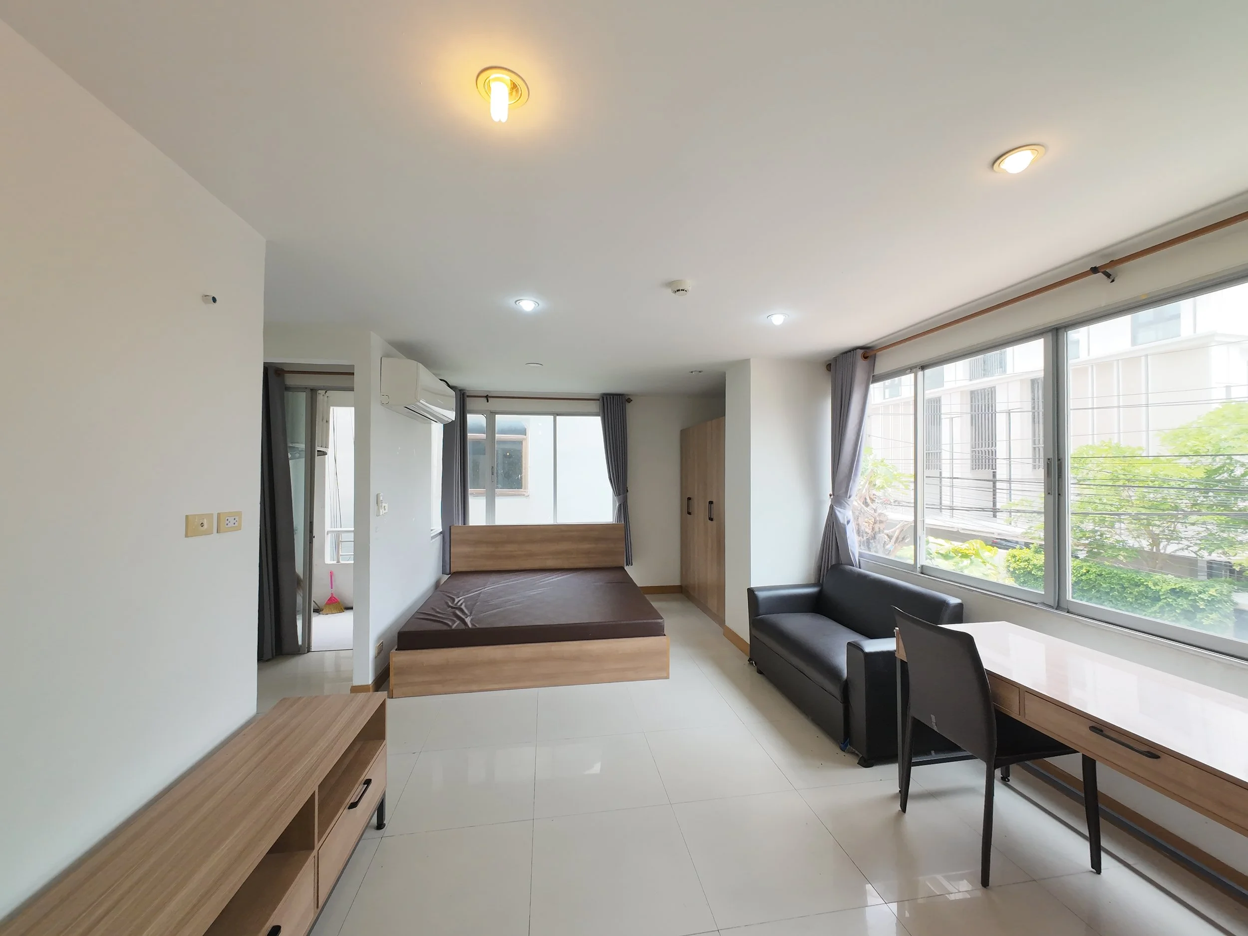 Modern studio apartment with a bed, black sofa, desk, large windows with gray curtains, white tiled floor, and minimalistic decor.