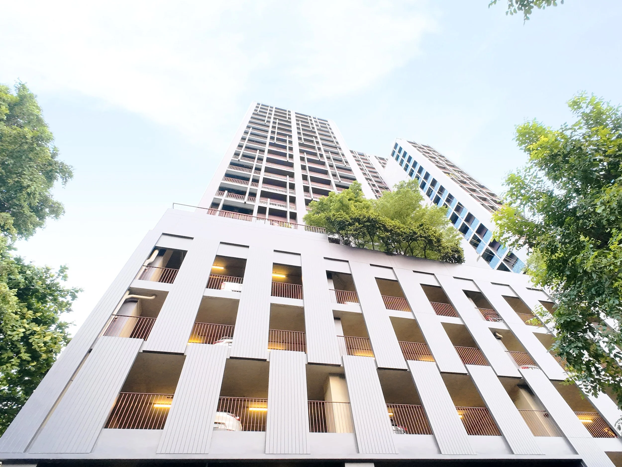 Low angle view of a modern high-rise building with white exterior and multiple balconies, surrounded by trees, against a bright blue sky.