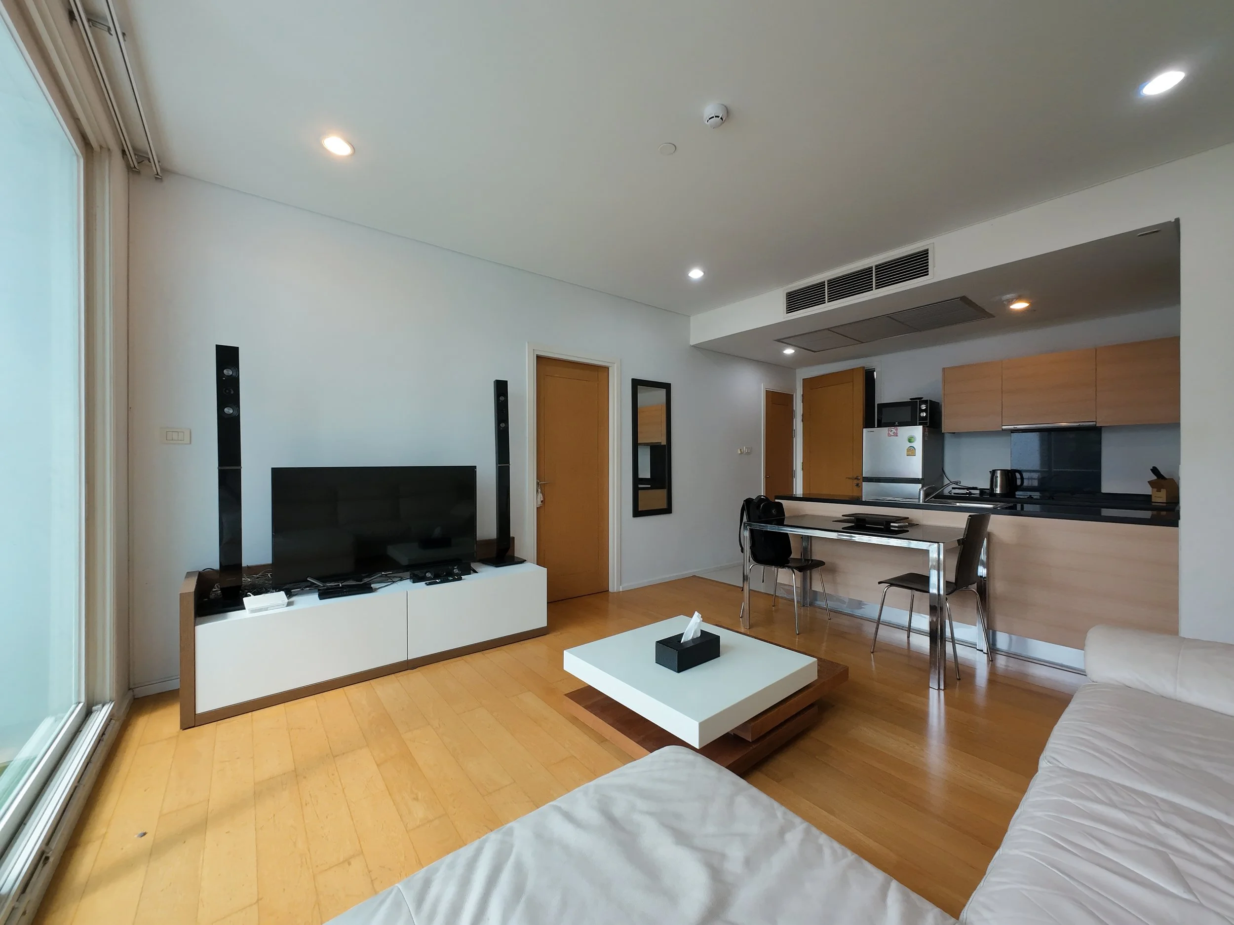 Living room with a flat-screen TV, tall speakers, small white coffee table, and a white sofa, with a view of the kitchen area that includes wooden cabinets, a refrigerator, microwave, and chairs at a dining table. Recessed lighting is on the ceiling, and there is a sliding glass door on the left.