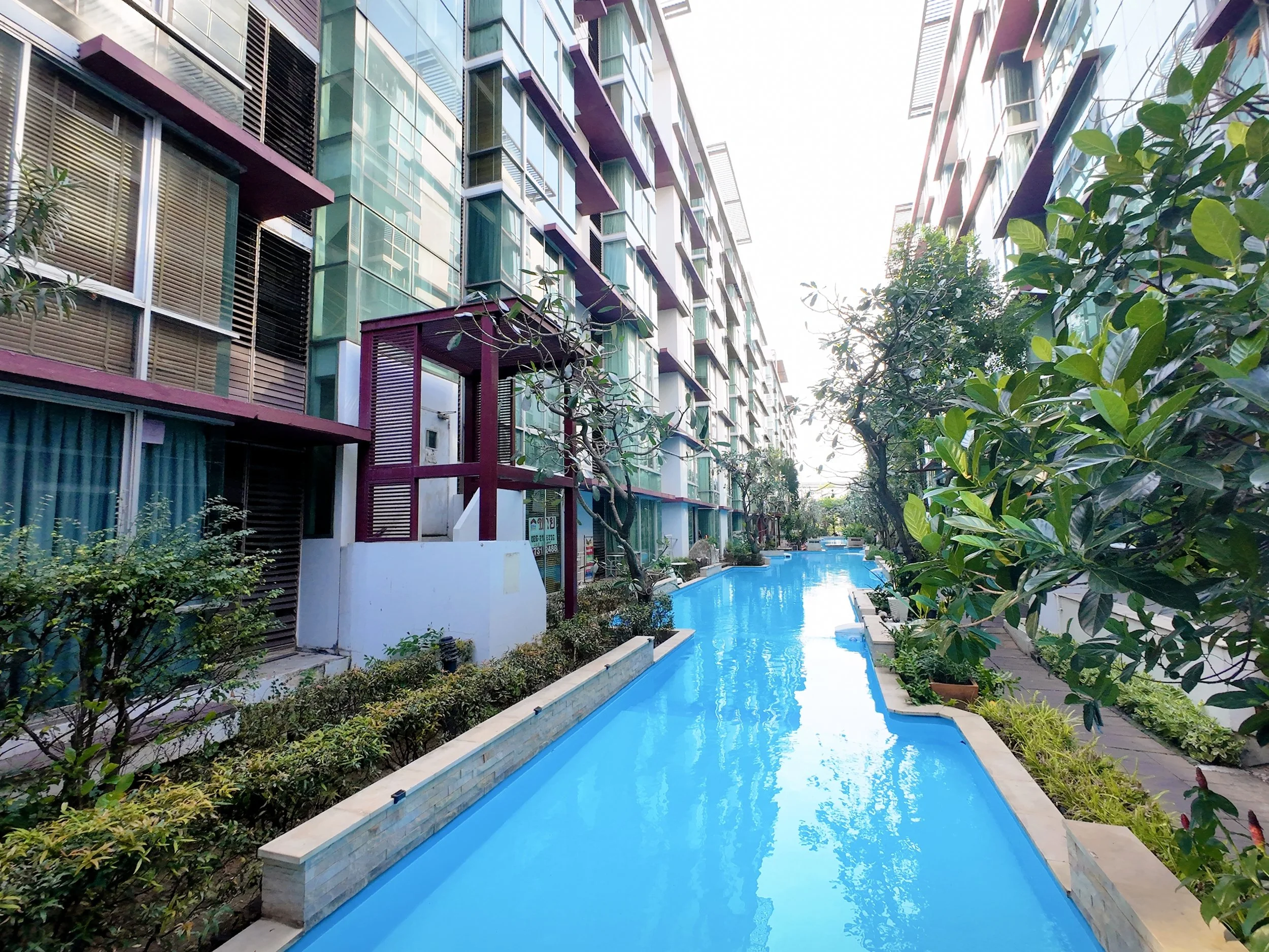 View of a modern apartment complex with a swimming pool running through the center, surrounded by greenery and multi-story glass buildings.