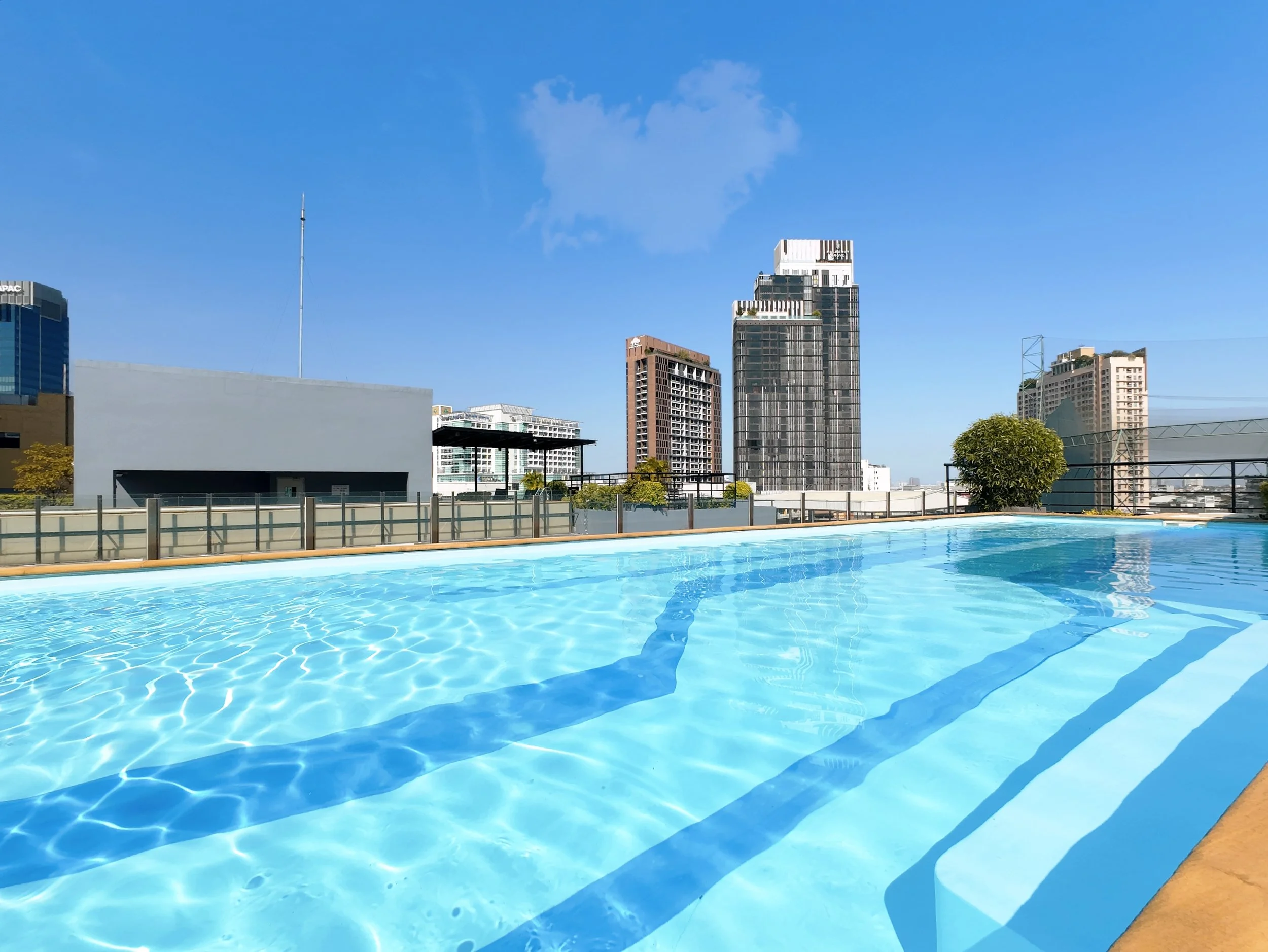 A rooftop swimming pool with a city skyline in the background, including modern high-rise buildings under a clear blue sky.
