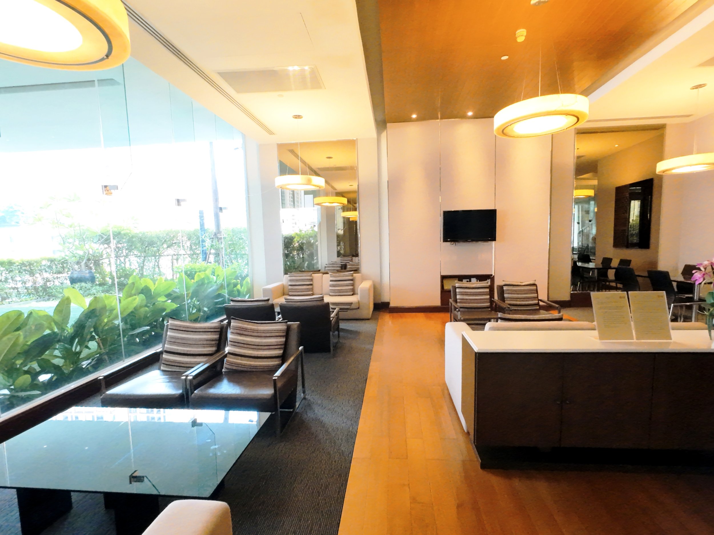 Indoor seating area with chairs and sofas, large glass window with views of greenery outside, modern lighting fixtures on the ceiling, and a flat-screen TV mounted on the wall.