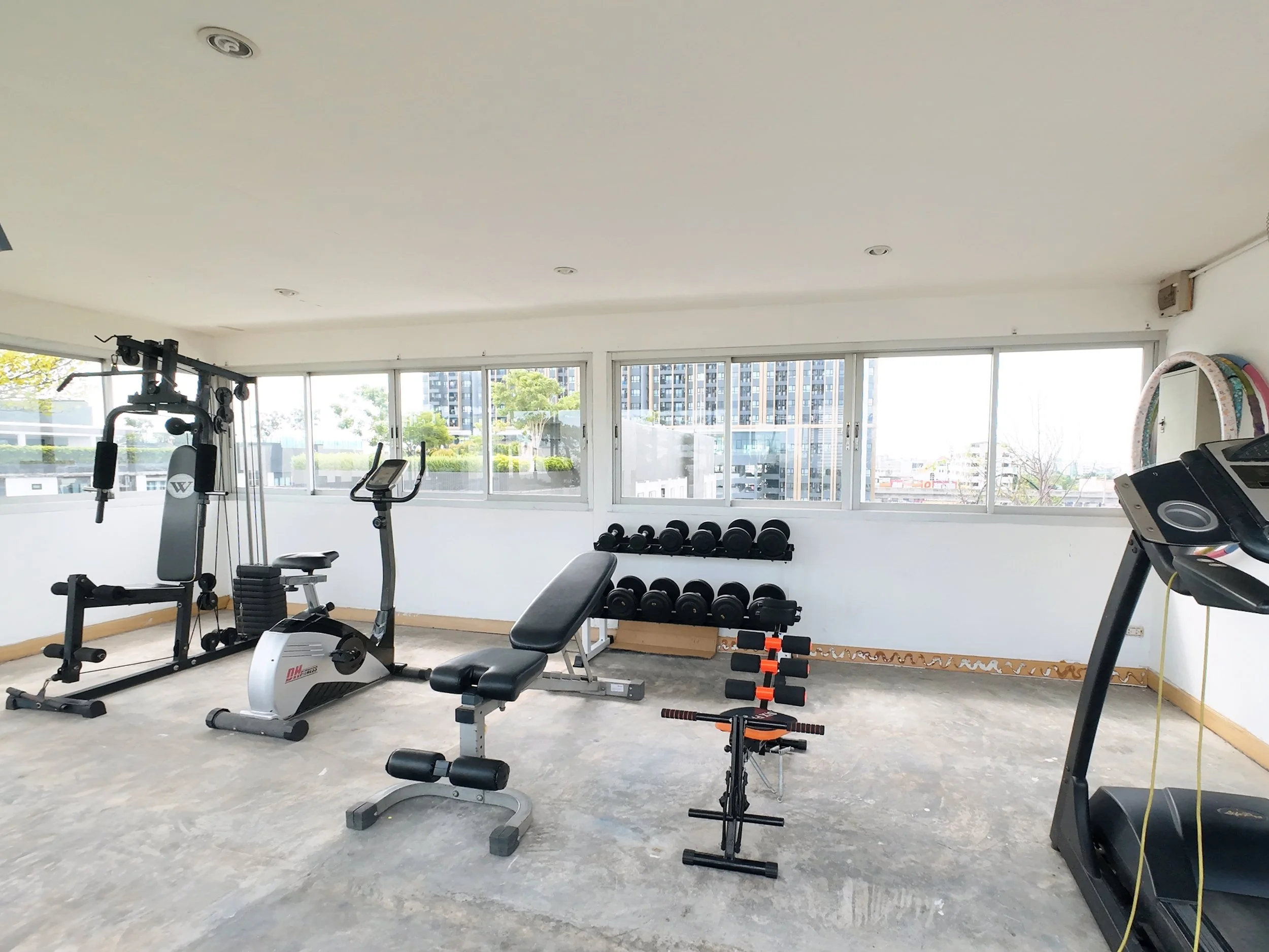 Empty gym room with exercise equipment including treadmill, elliptical, weight bench, dumbbells, and other fitness machines, with large windows revealing tall buildings and greenery outside.