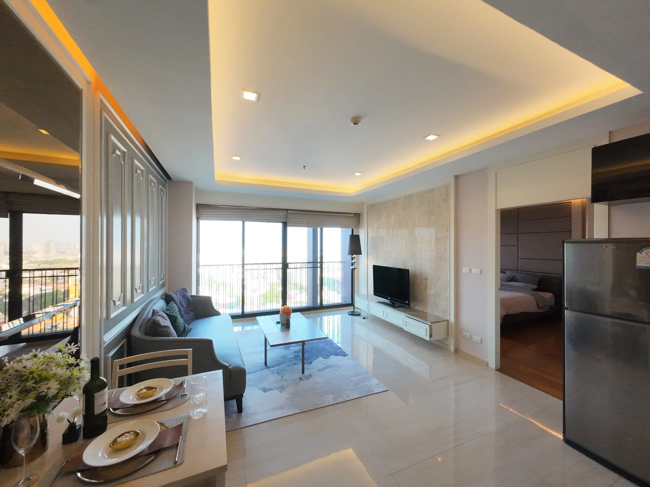A modern living room with a sofa, coffee table, TV, and large windows overlooking a cityscape during daytime.