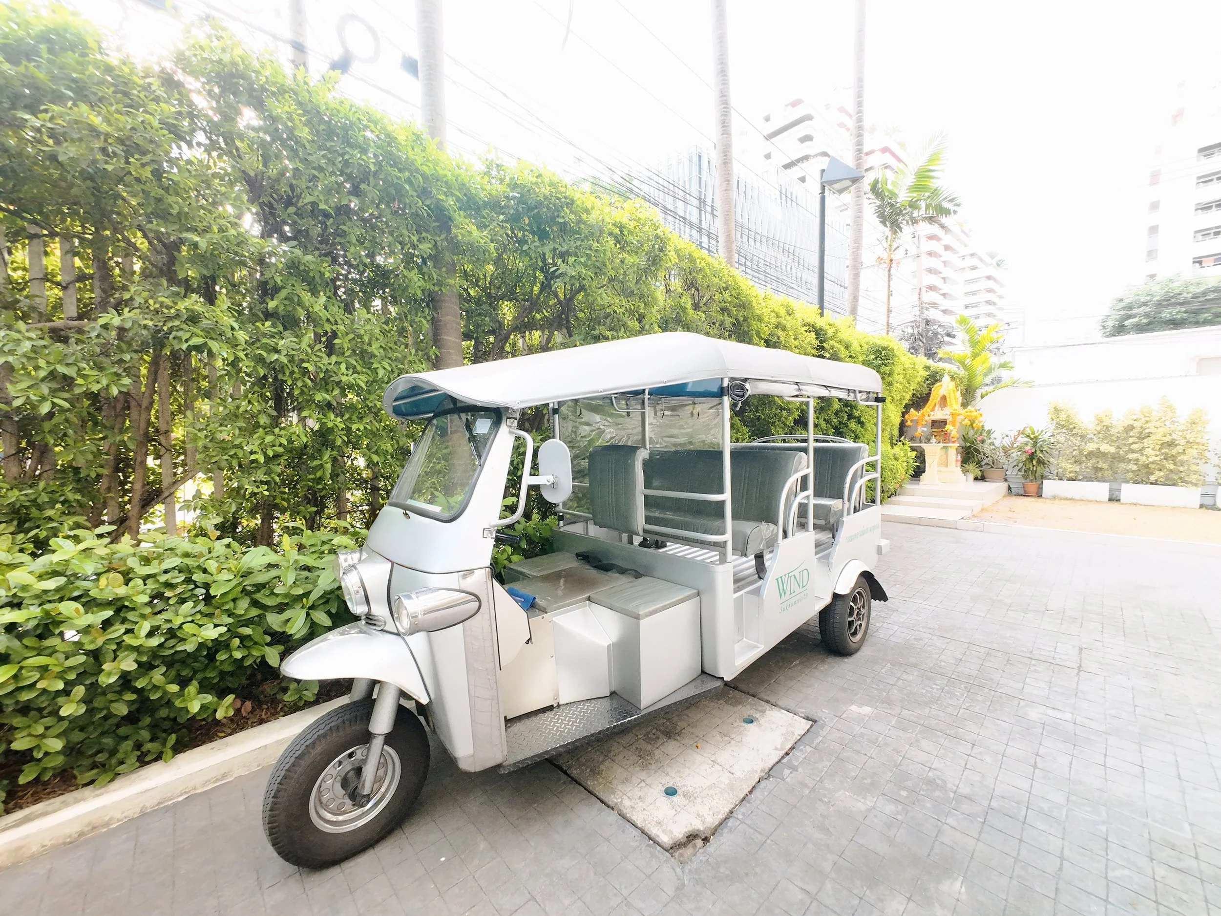 A white electric golf cart with seating for several passengers, parked on a paved sidewalk near a green hedge and trees with urban buildings in the background.