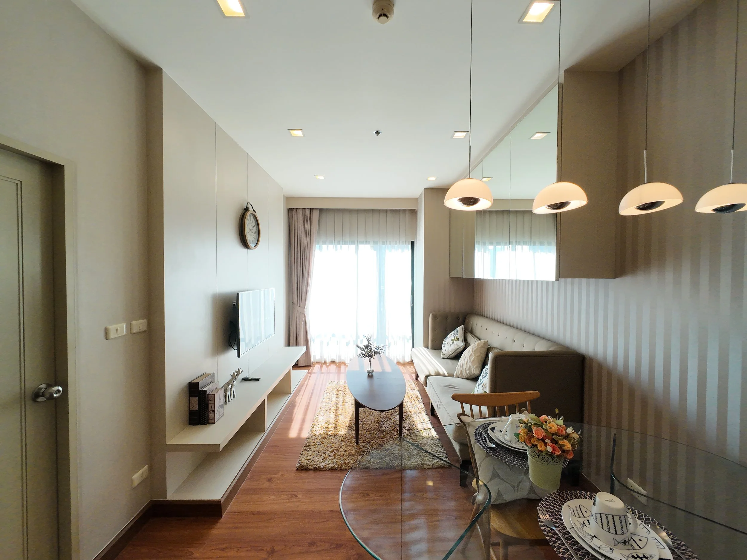 Living room with beige sofa, glass coffee table, wall-mounted TV, striped wallpaper, curtains, pendant lights, and a round wall clock.