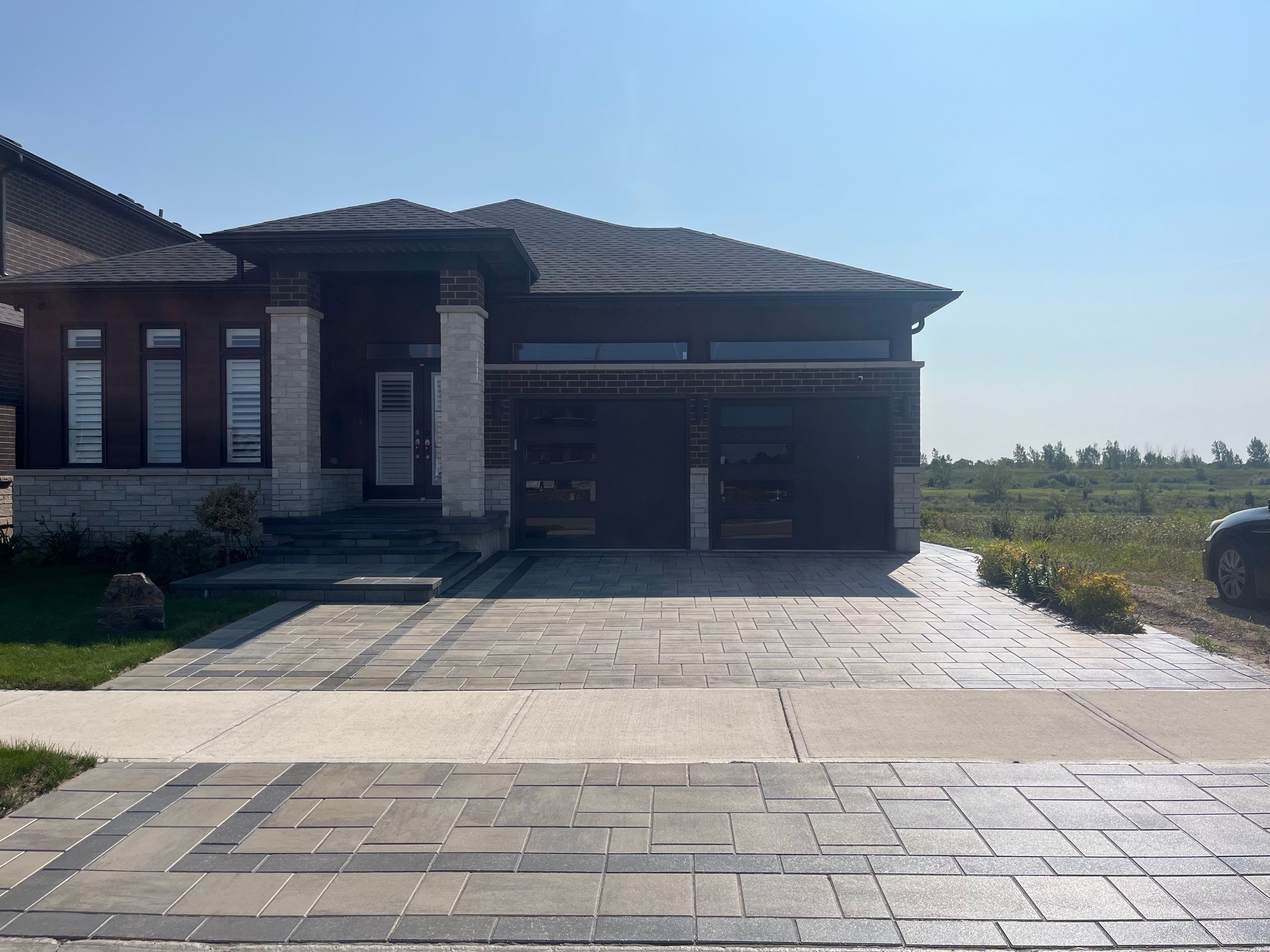 A paved outdoor area with a pattern of light and dark gray rectangular tiles, bordered by darker gray tiles, and surrounded by green bushes and trees.