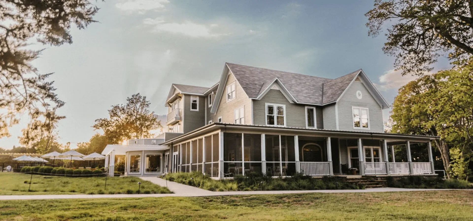 A large, multi-story house with a screened-in porch, surrounded by trees and a grassy lawn, under a partly cloudy sky.