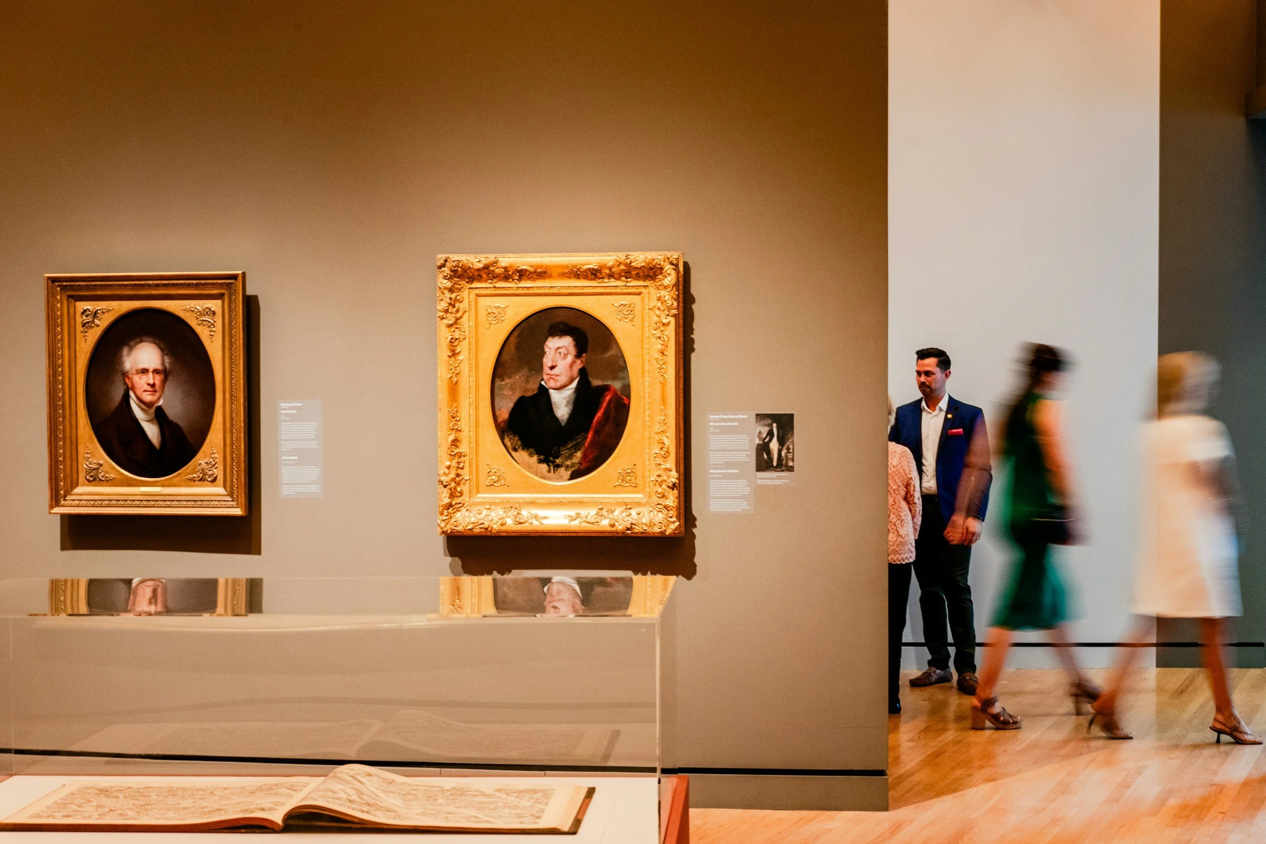 People viewing classical portrait paintings in a museum, with one man in a dark suit and three women walking past.