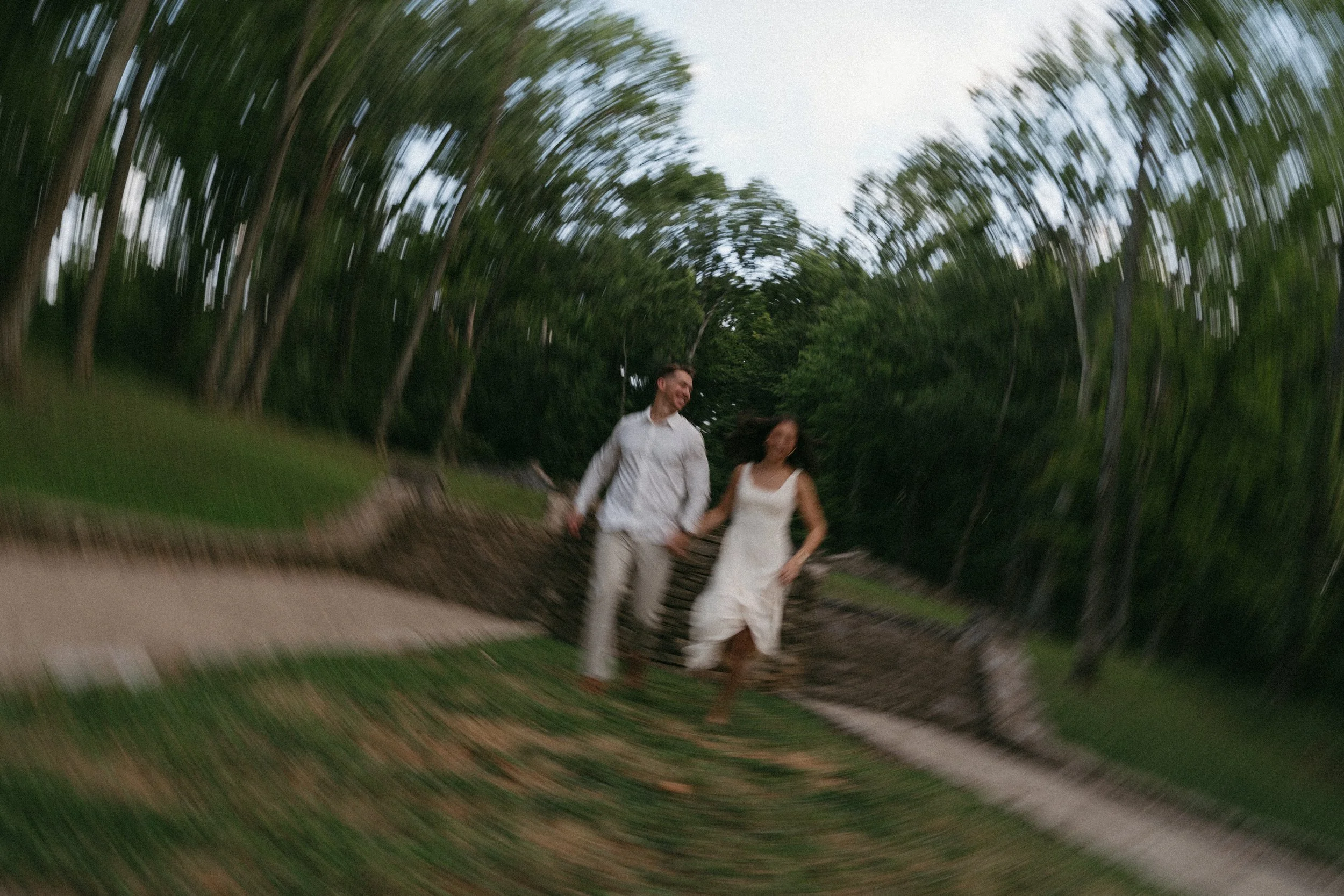 A couple walking hand in hand outdoors on a trail, smiling and looking at each other, surrounded by trees and greenery.
